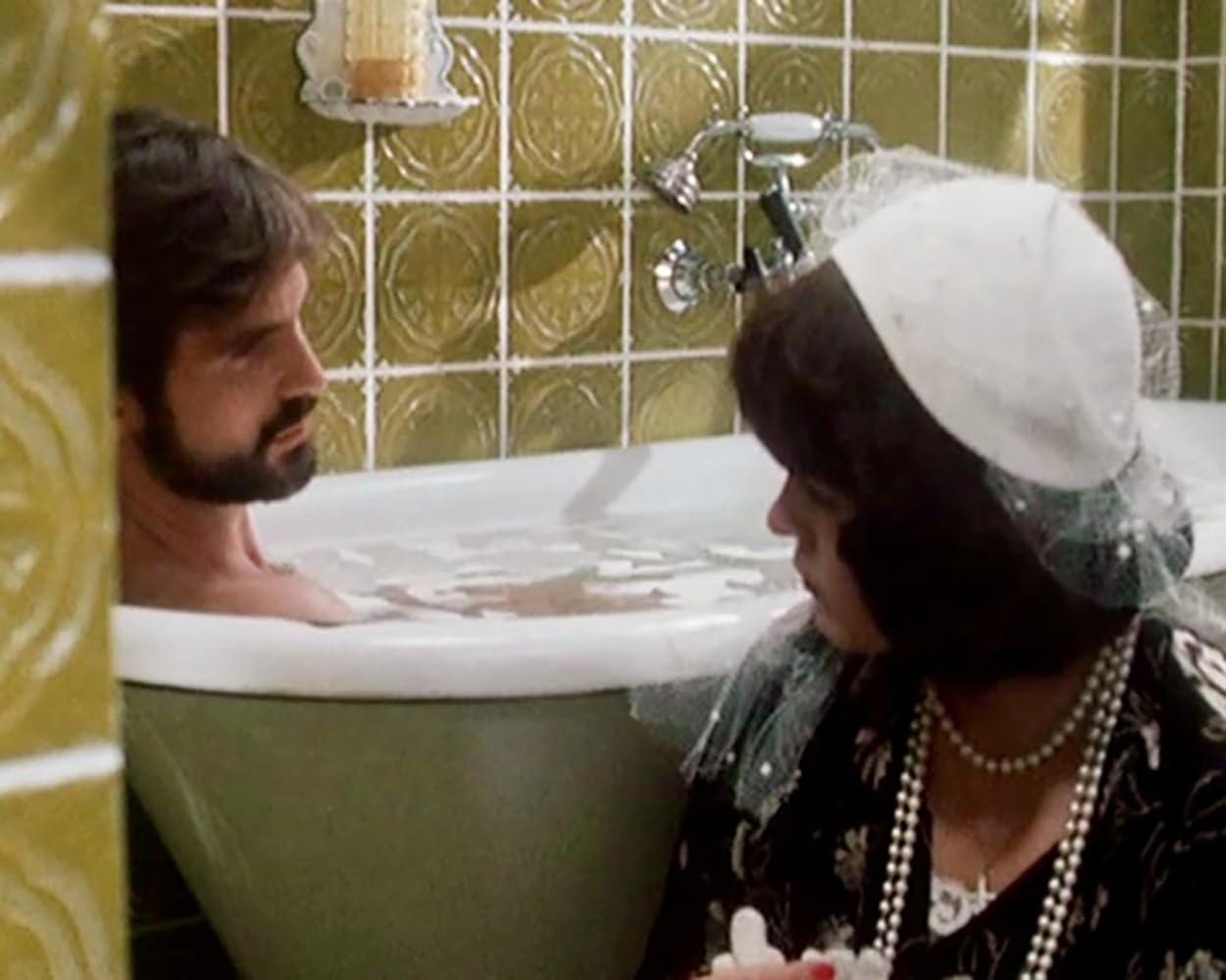 Ein bärtiger Mann in einer Badewanne schaut eine Frau mit weißem Hut und Perlen an.