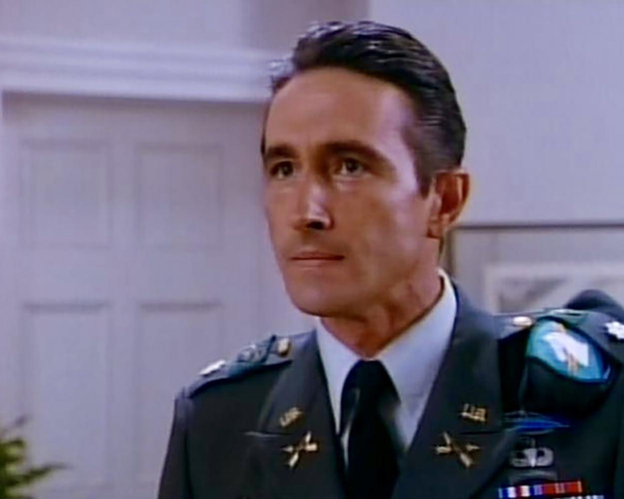 Ein Soldat in Uniform mit ernstem Gesichtsausdruck vor weißer Tür.
