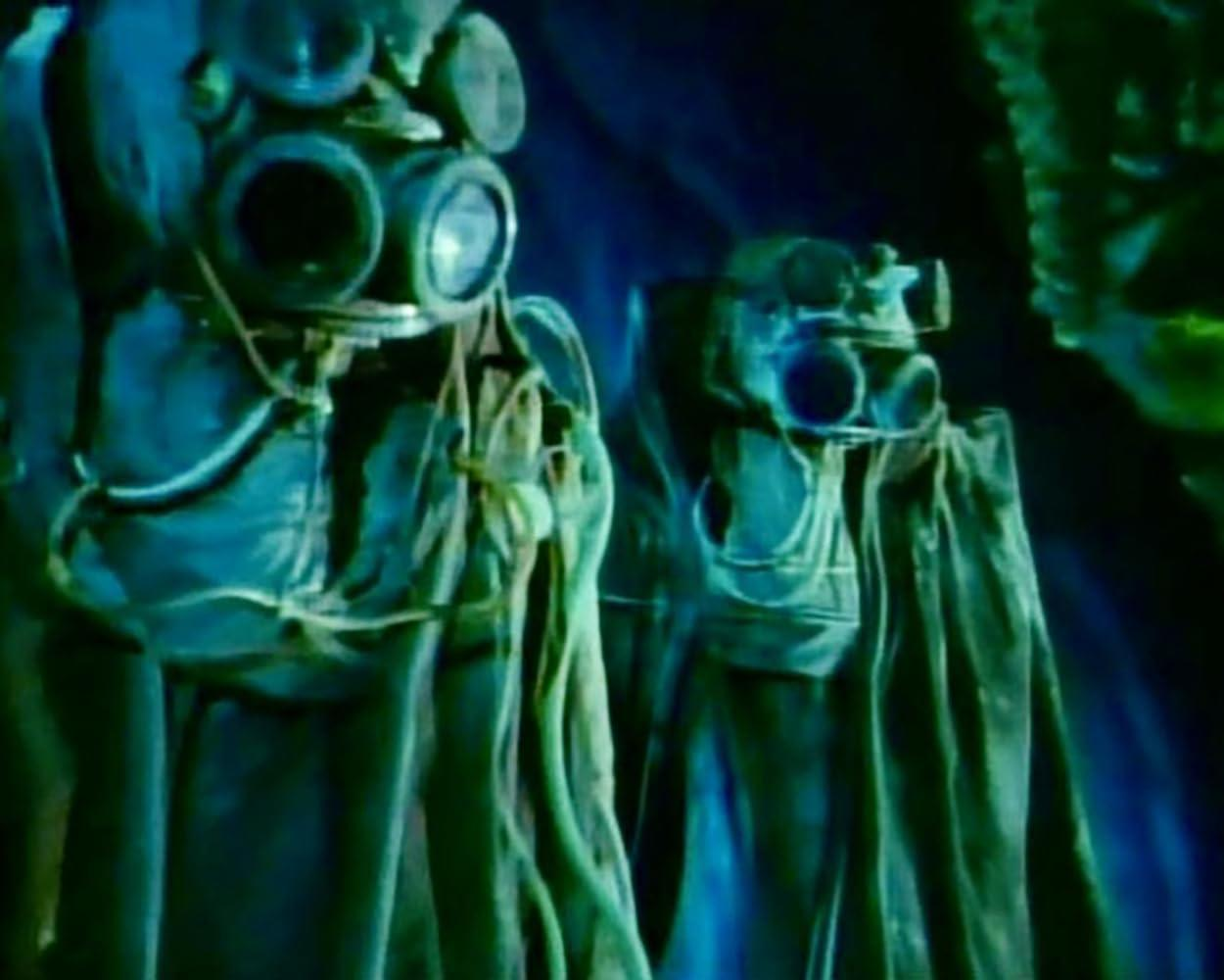 Zwei Figuren in dunkler Kleidung und mit großen Gasmasken stehen in einem blau beleuchteten Raum.