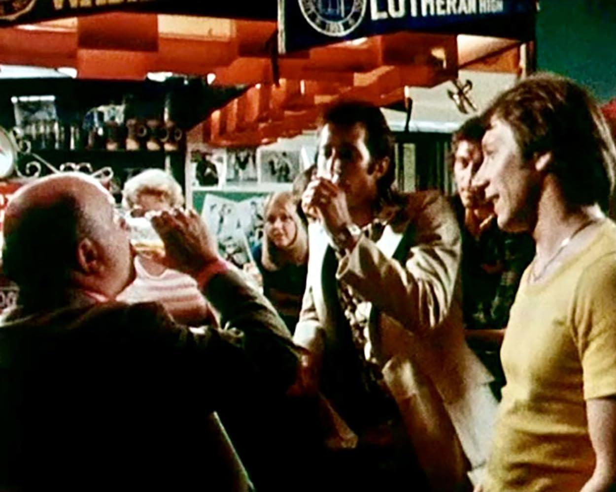 Menschen trinken und unterhalten sich in einer lebhaften Bar, im Hintergrund hängen Regale und Plakate.