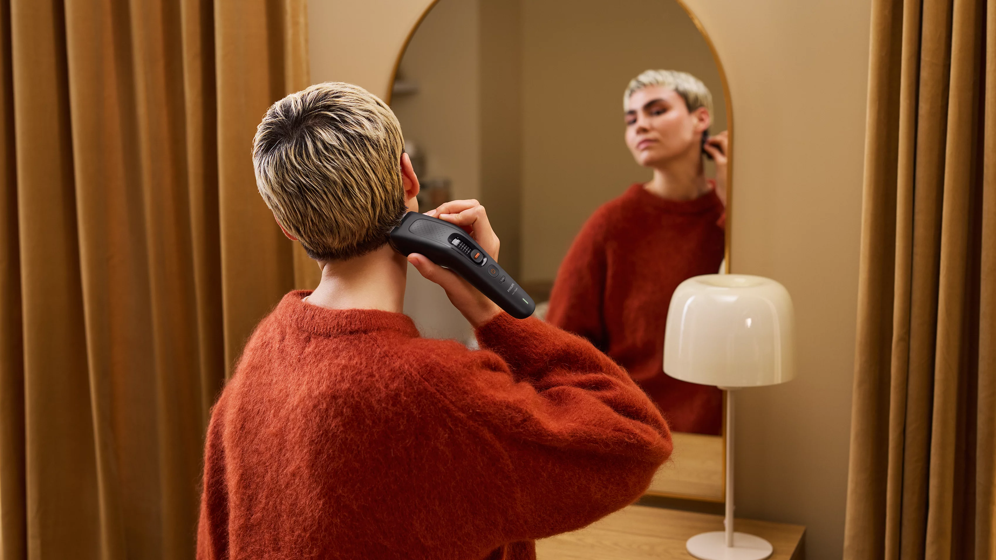 Eine Person mit rotem Pullover rasiert sich im Spiegel die Haare mit einem Haarschneider.
