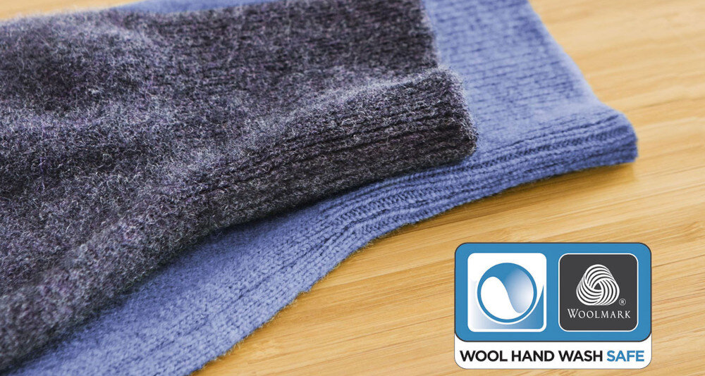 Dwa złożone swetry w ciemnym i jasnym odcieniu niebieskiego na drewnianej powierzchni z logo Woolmark do prania ręcznego.