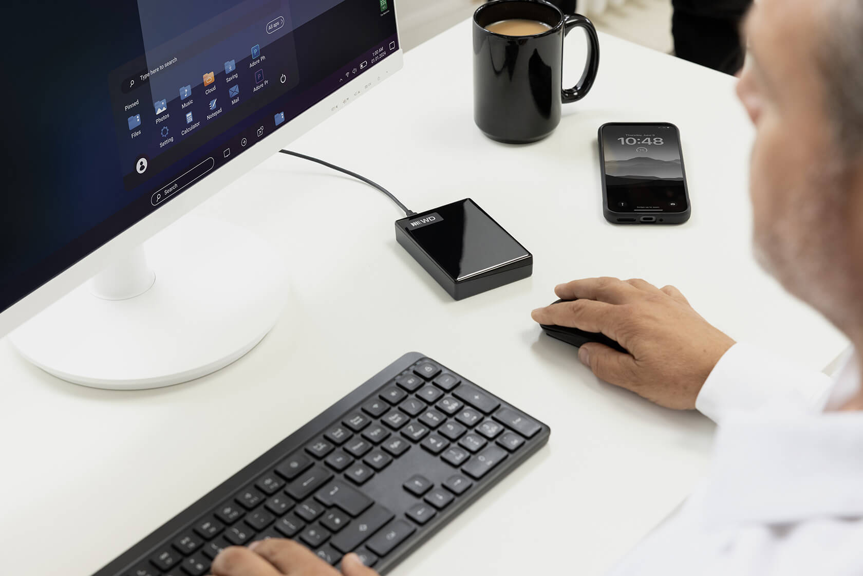 Ein Mann nutzt einen Computer an einem weißen Schreibtisch mit externer Festplatte, Tastatur, Maus, Smartphone und Tasse.