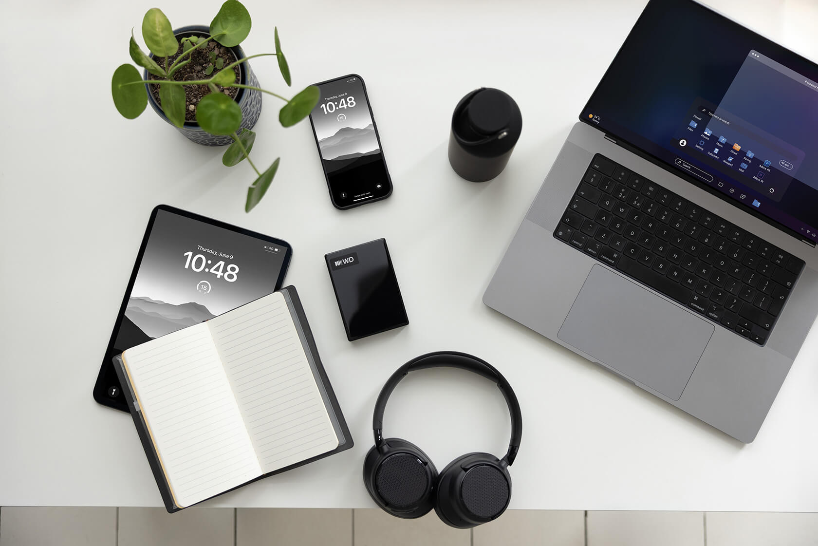 Ein weißer Schreibtisch mit Laptop, Tablet, Smartphone, Notizbuch, Kopfhörern, WD-Festplatte und einer Pflanze.