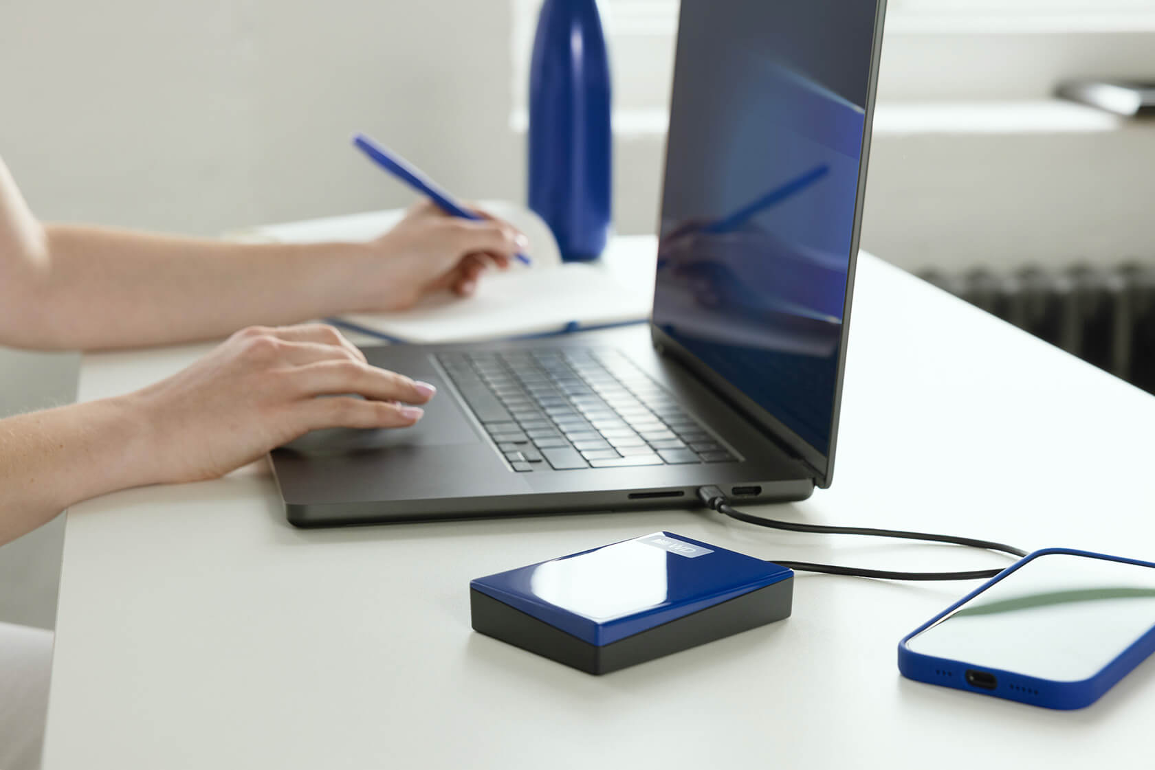Person arbeitet am Laptop an einem weißen Tisch mit externer Festplatte und Telefon.