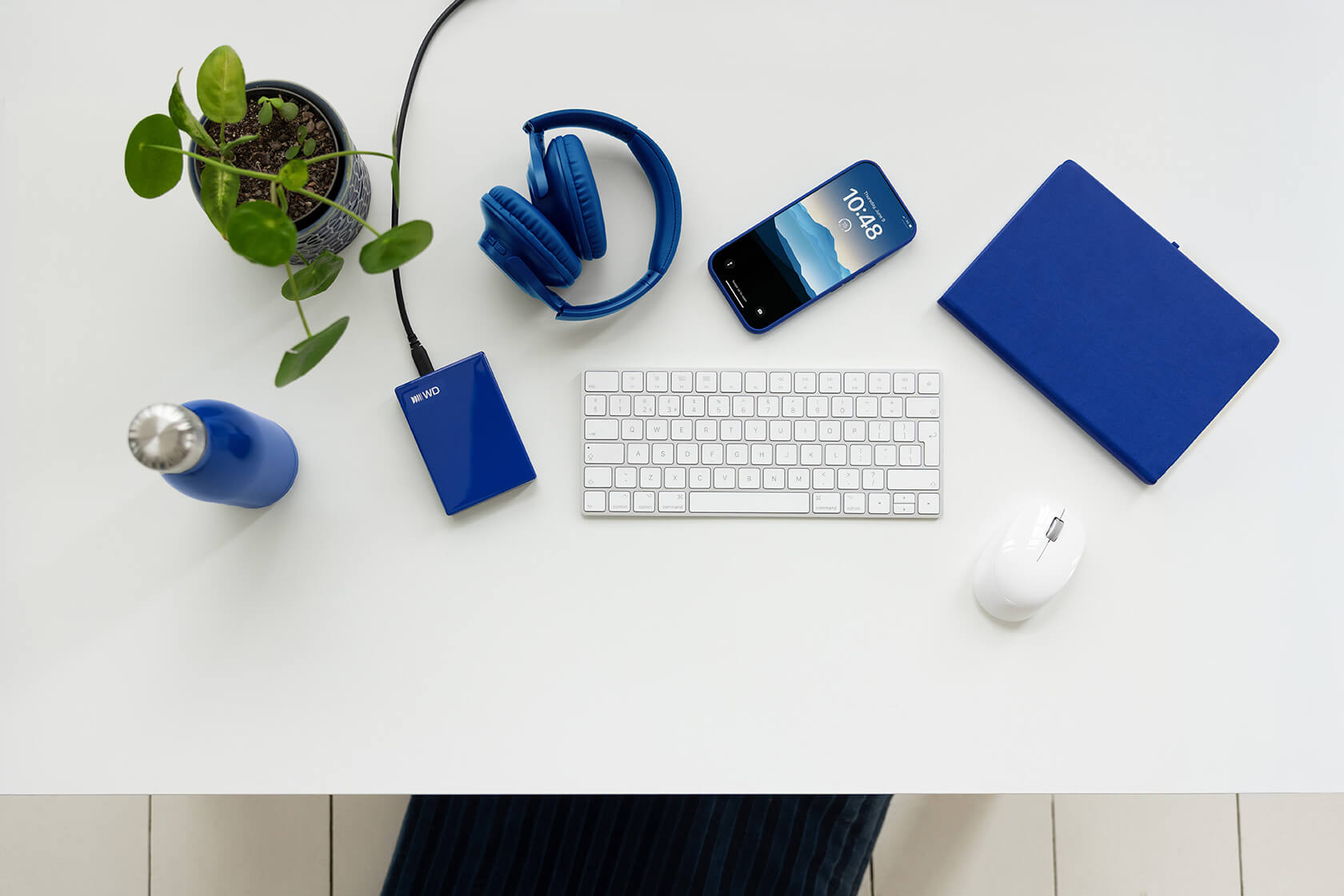 Ein weißer Schreibtisch mit blauen Accessoires: Kopfhörer, Smartphone, Notizbuch, Festplatte, Flasche und Pflanze.