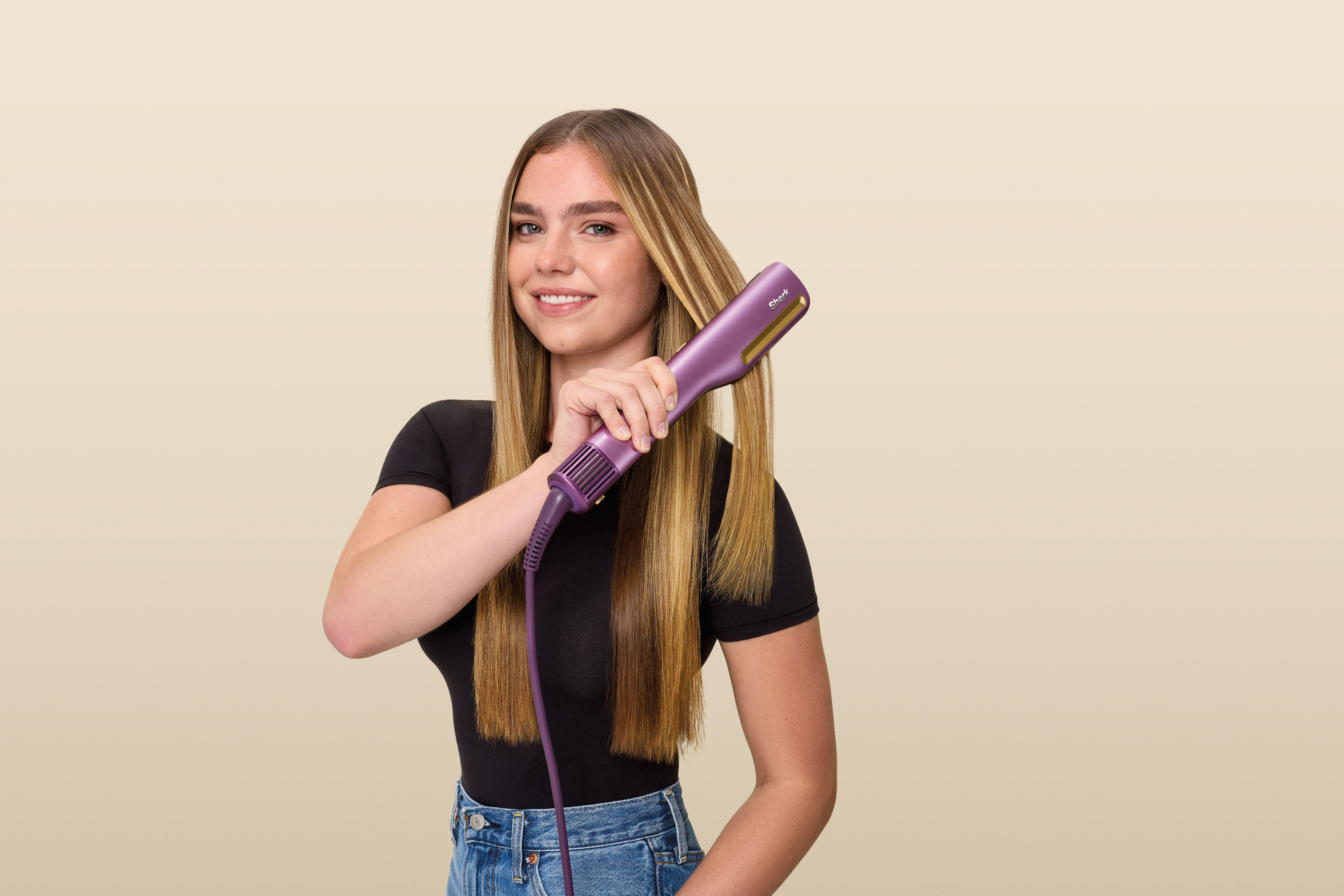 Eine lächelnde Frau hält einen violetten Haarglätter und stylt ihr langes blondes Haar vor einem beigen Hintergrund.