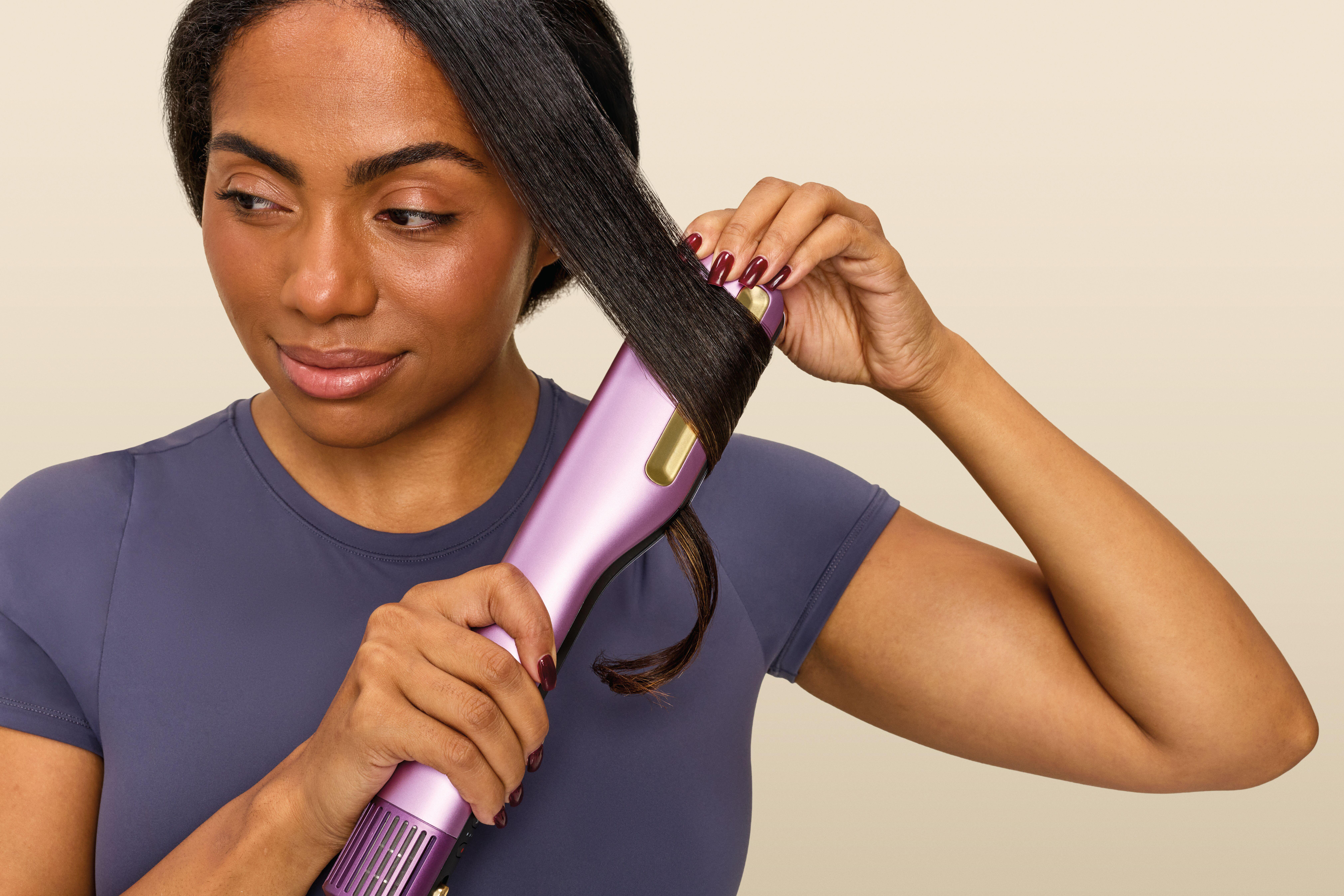 Eine Frau stylt ihr dunkles Haar mit einem pink-goldenen Lockenstab vor neutralem Hintergrund.