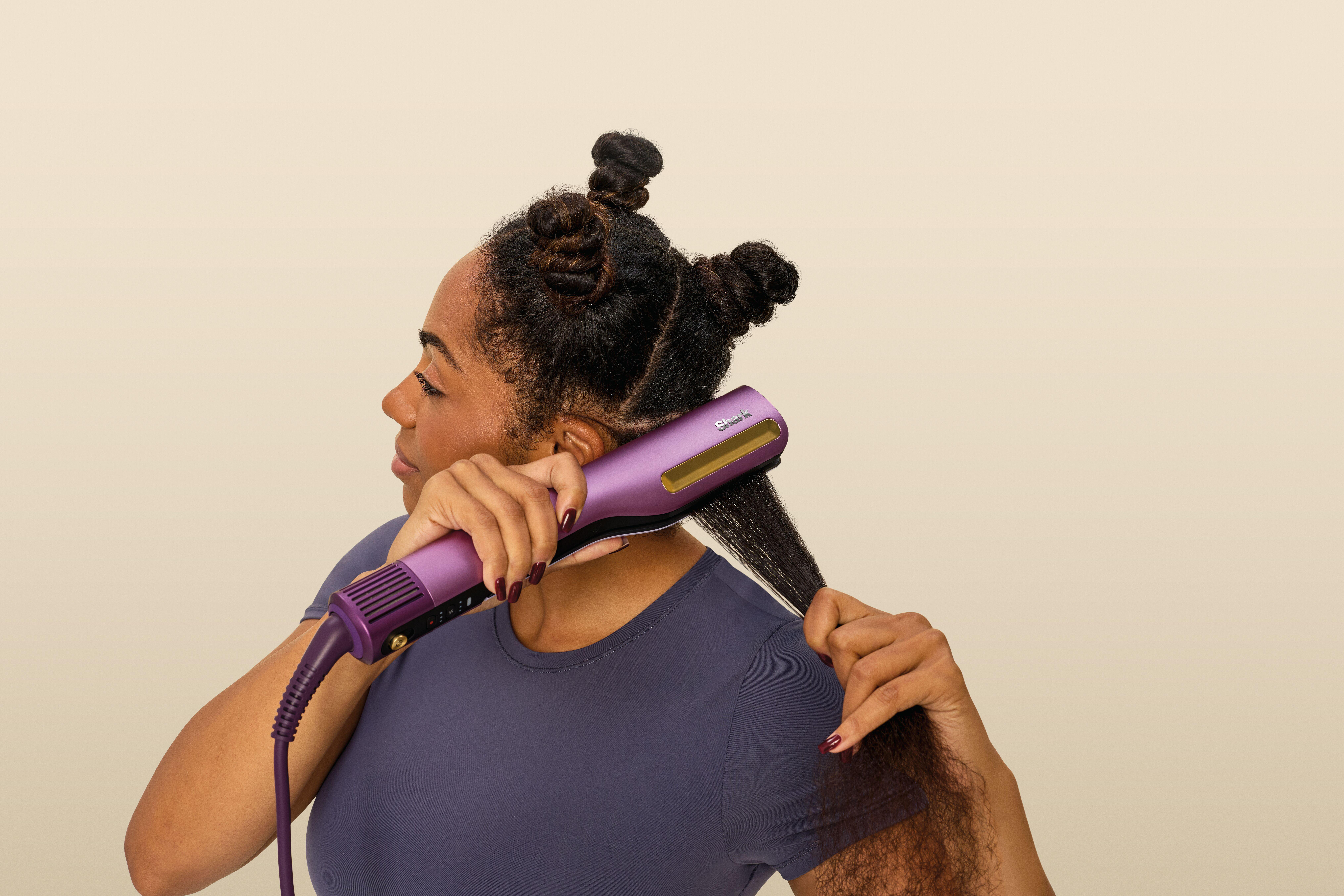 Eine Frau mit drei kleinen Haarknoten glättet ihr langes Haar mit einem violetten Glätteisen vor neutralem Hintergrund.