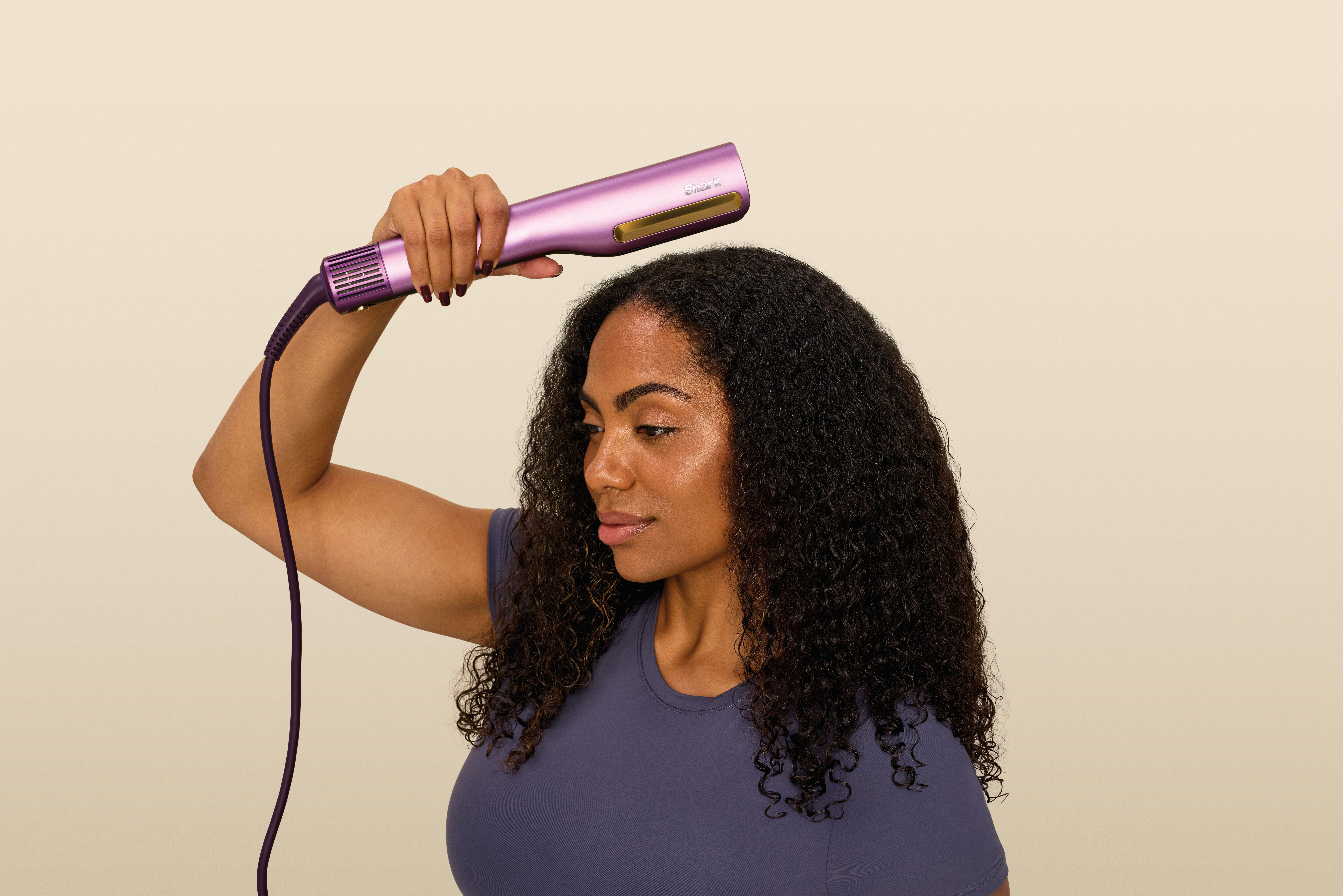 Eine Frau hält ein lilafarbenes Stylinggerät in der Hand, bereit, ihre dunklen Locken zu glätten.