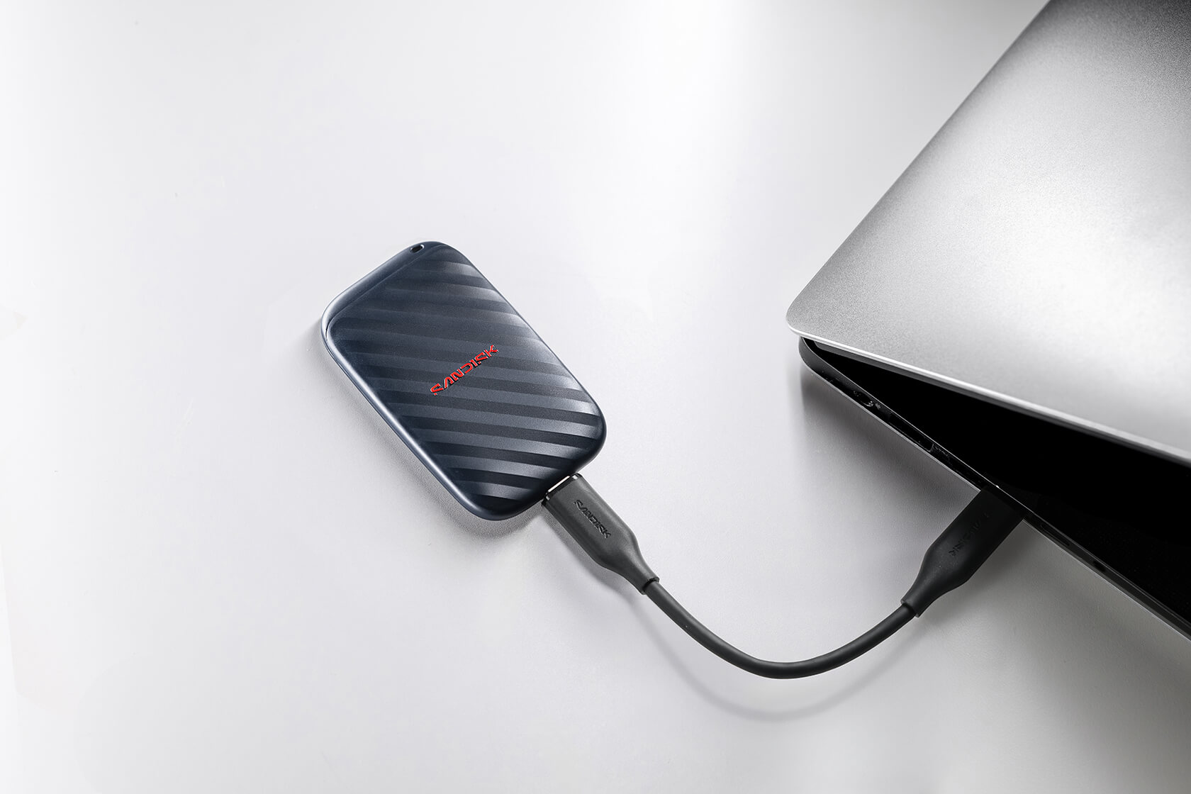 Eine schwarze SanDisk SSD ist über ein Kabel an einen silbernen Laptop auf weißem Tisch angeschlossen.