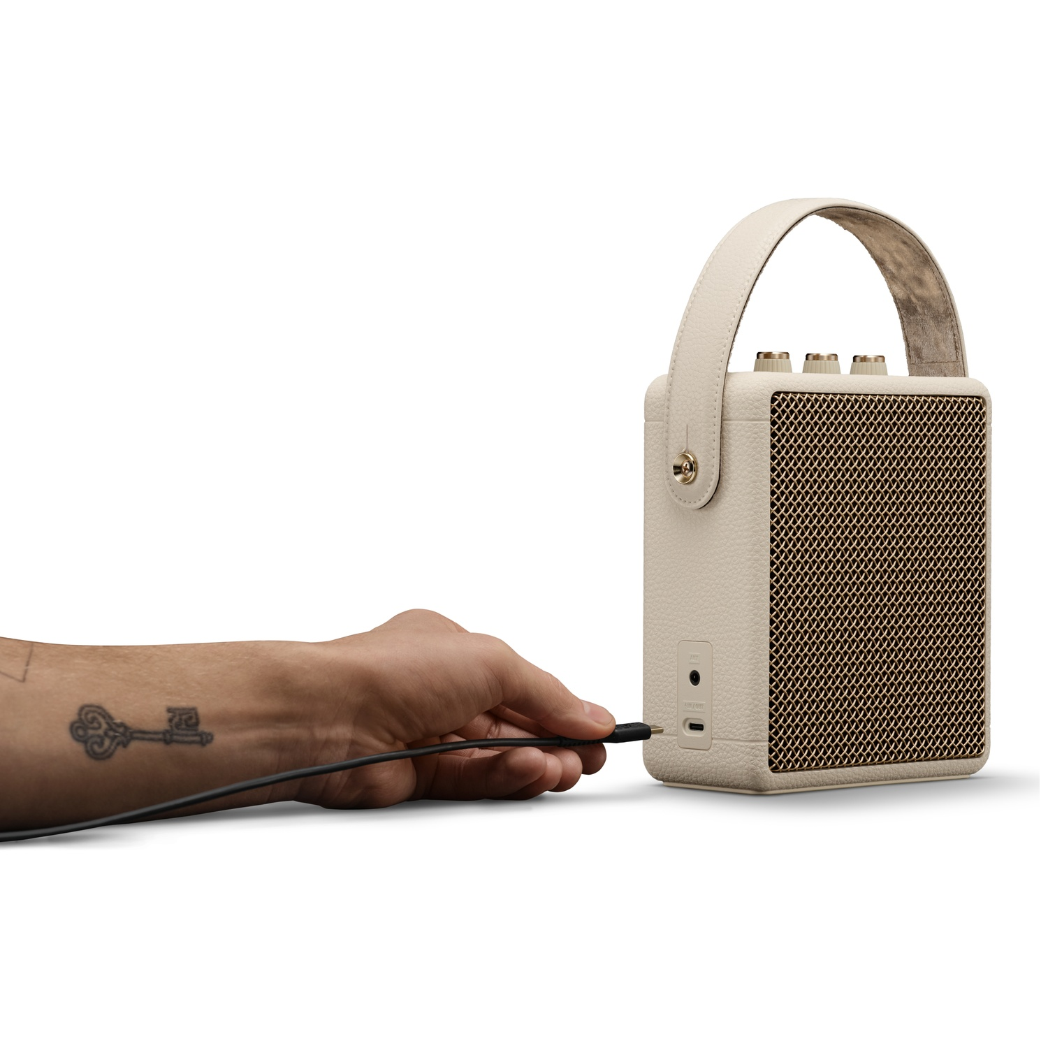 A tattooed hand plugging a black cable into a cream-colored portable speaker on a white background.