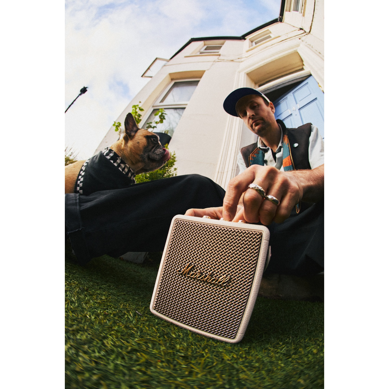 A man in a cap adjusts a Marshall speaker while a dog sits on grass in front of a white house.