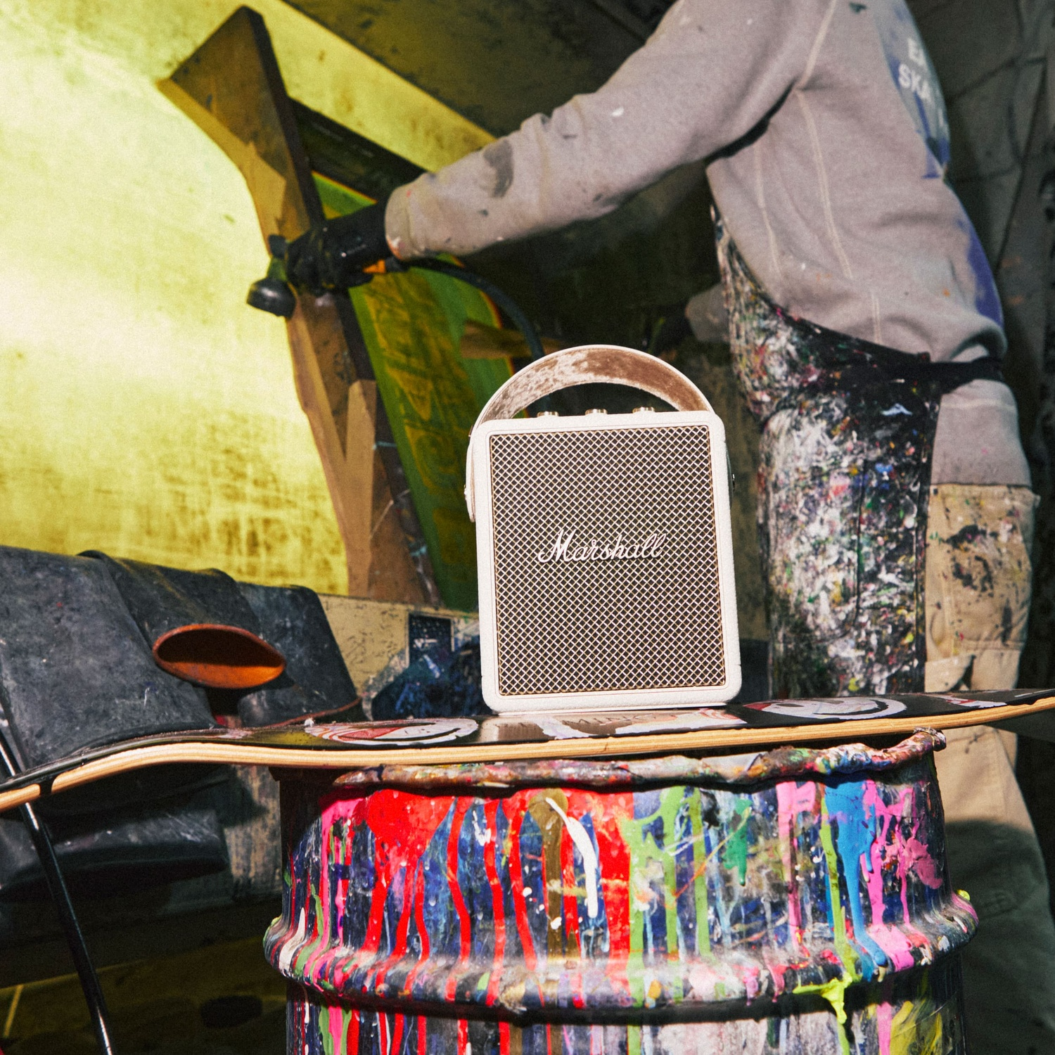 A white Marshall speaker sits on a paint-splattered drum while an artist works in the blurry background.