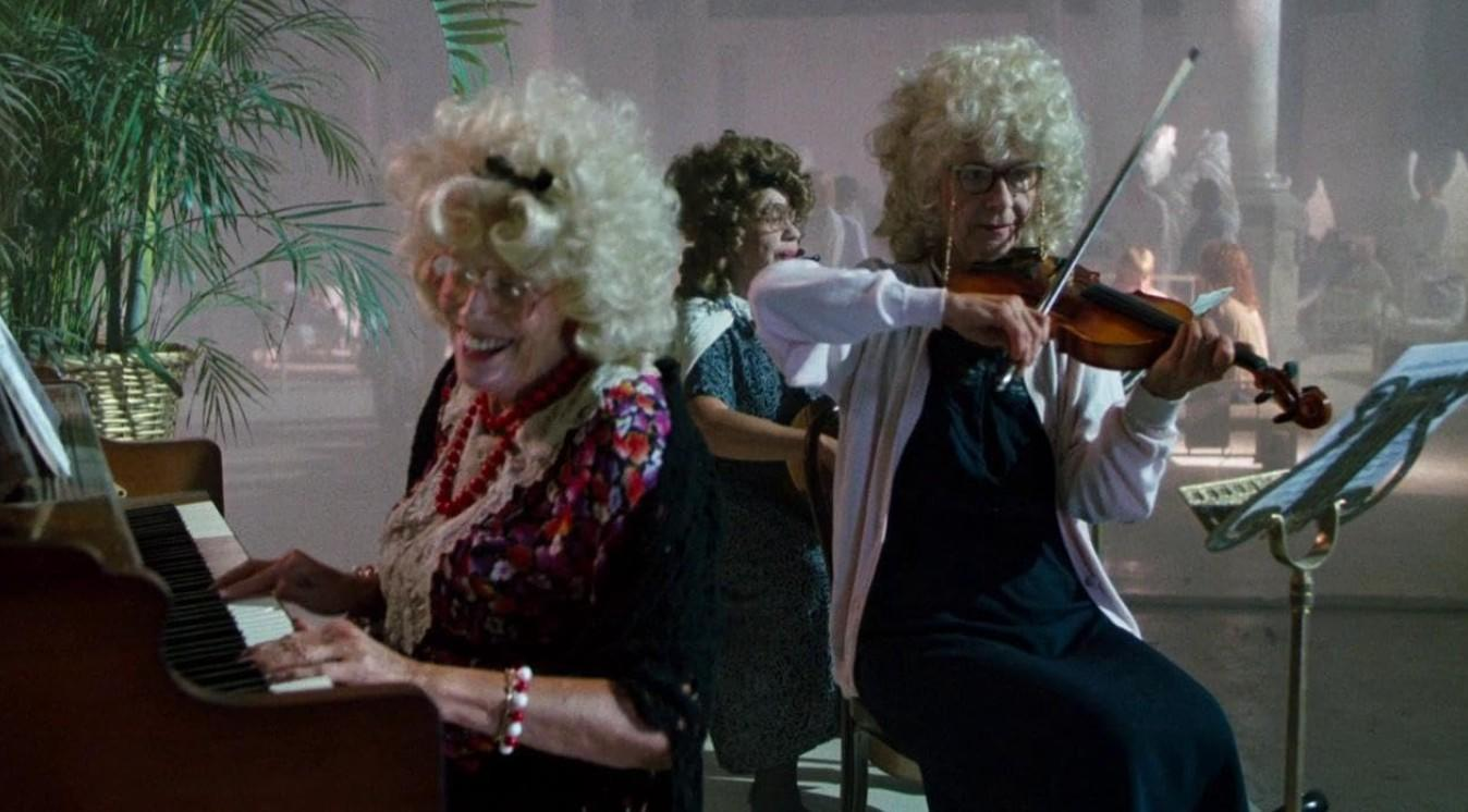 Ältere Frauen in blonden Perücken spielen in einem Raum Klavier und Geige.