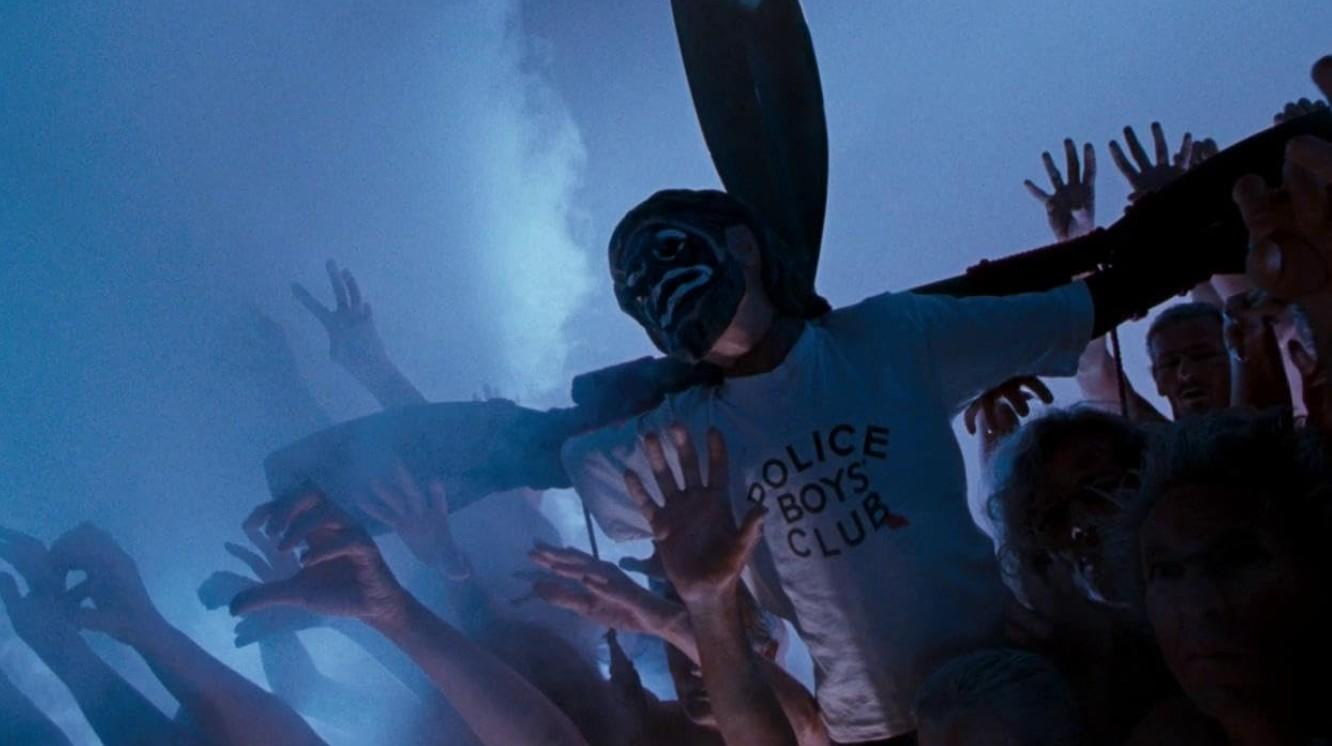 Eine maskierte Person in einem Police Boys Club T-Shirt ist von erhobenen Händen umgeben.