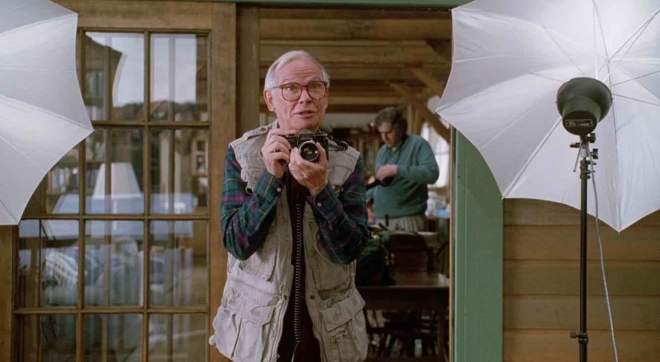 Ein älterer Mann mit Brille und Weste fotografiert in einem Raum mit Studiobeleuchtung.