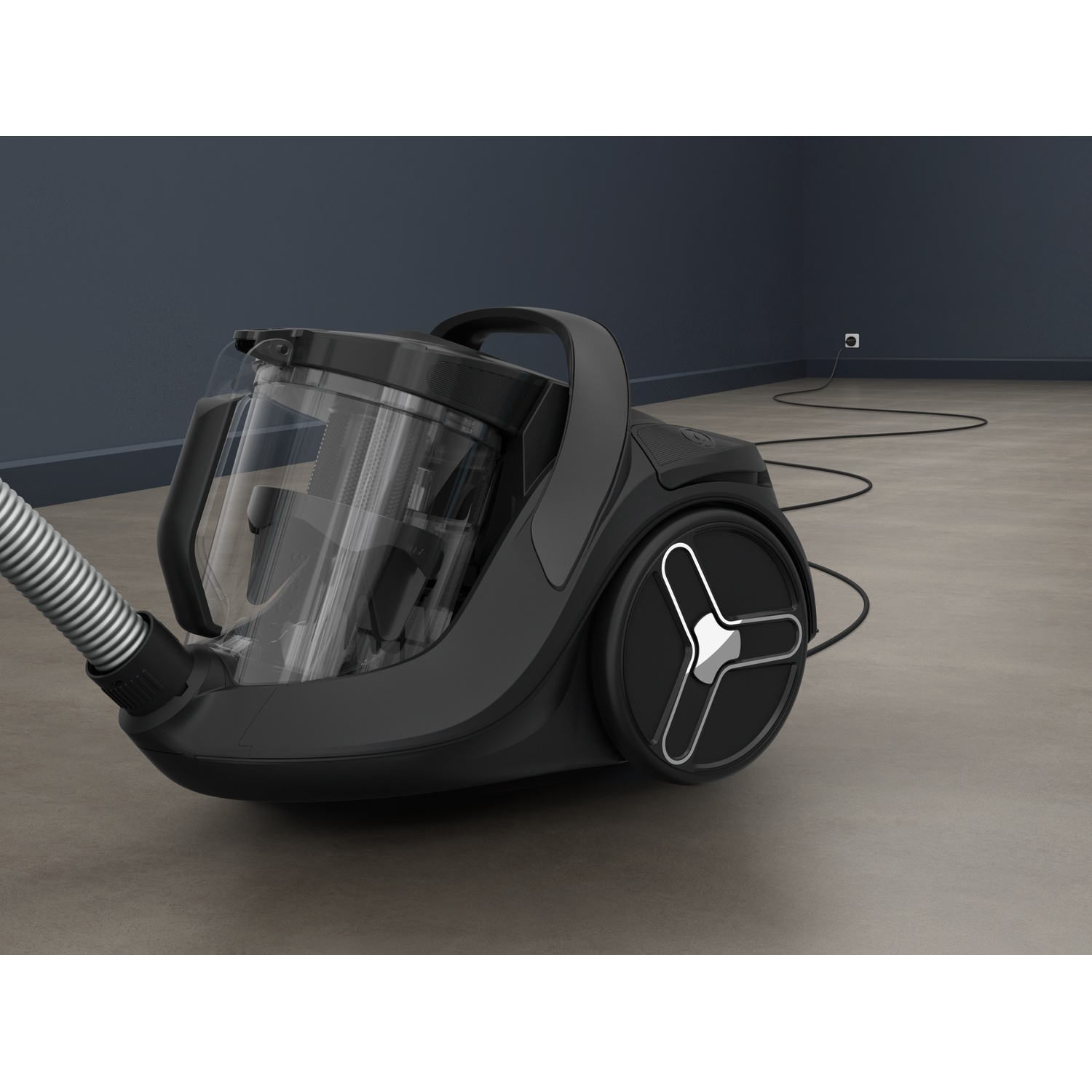 A black canister vacuum cleaner on a grey floor against a dark wall, with its cord plugged into a socket.