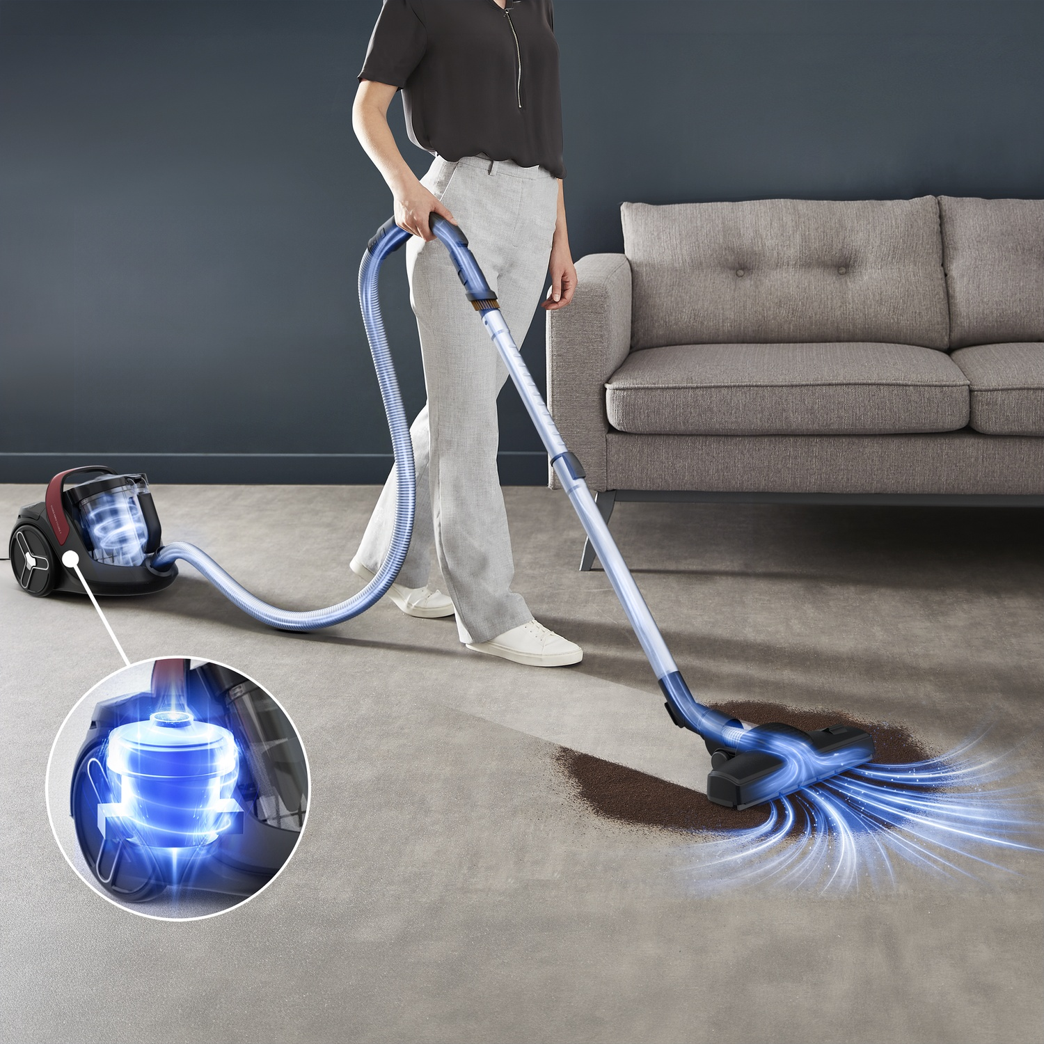 A woman vacuums dirt with a blue vacuum cleaner in front of a gray sofa.
