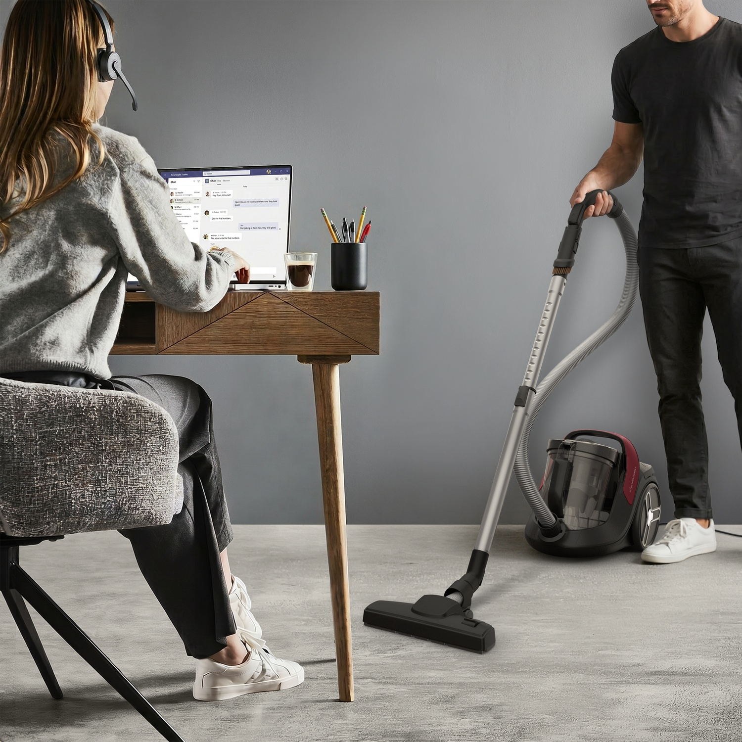 A woman sits at a wooden desk with a laptop, while a man vacuums the floor beside her.