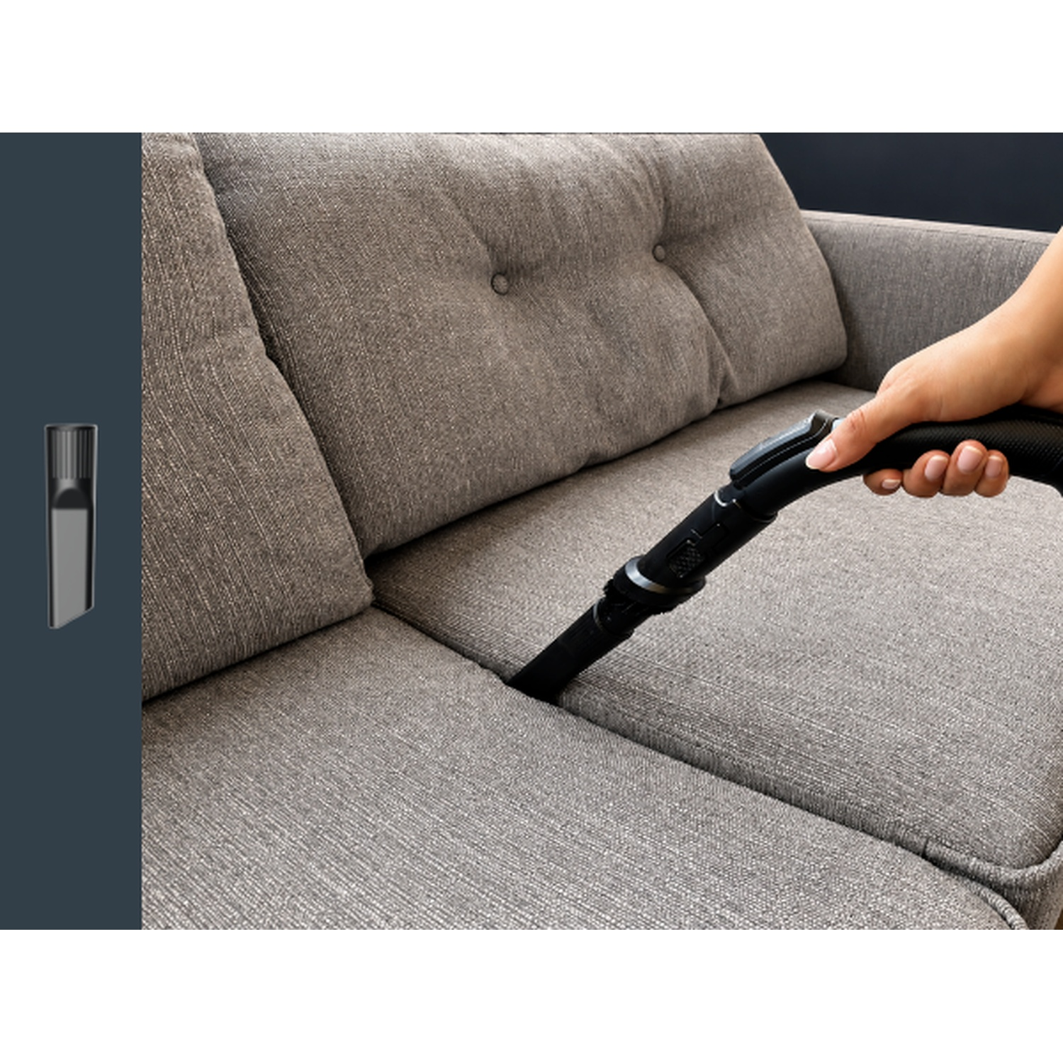 A hand uses a black attachment to vacuum the crevice of a gray fabric sofa.