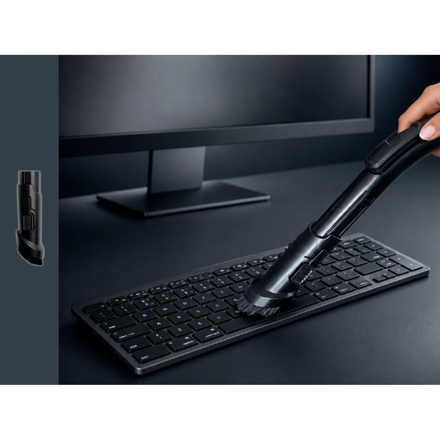 A hand cleans a black computer keyboard with a small black vacuum attachment on the desk.