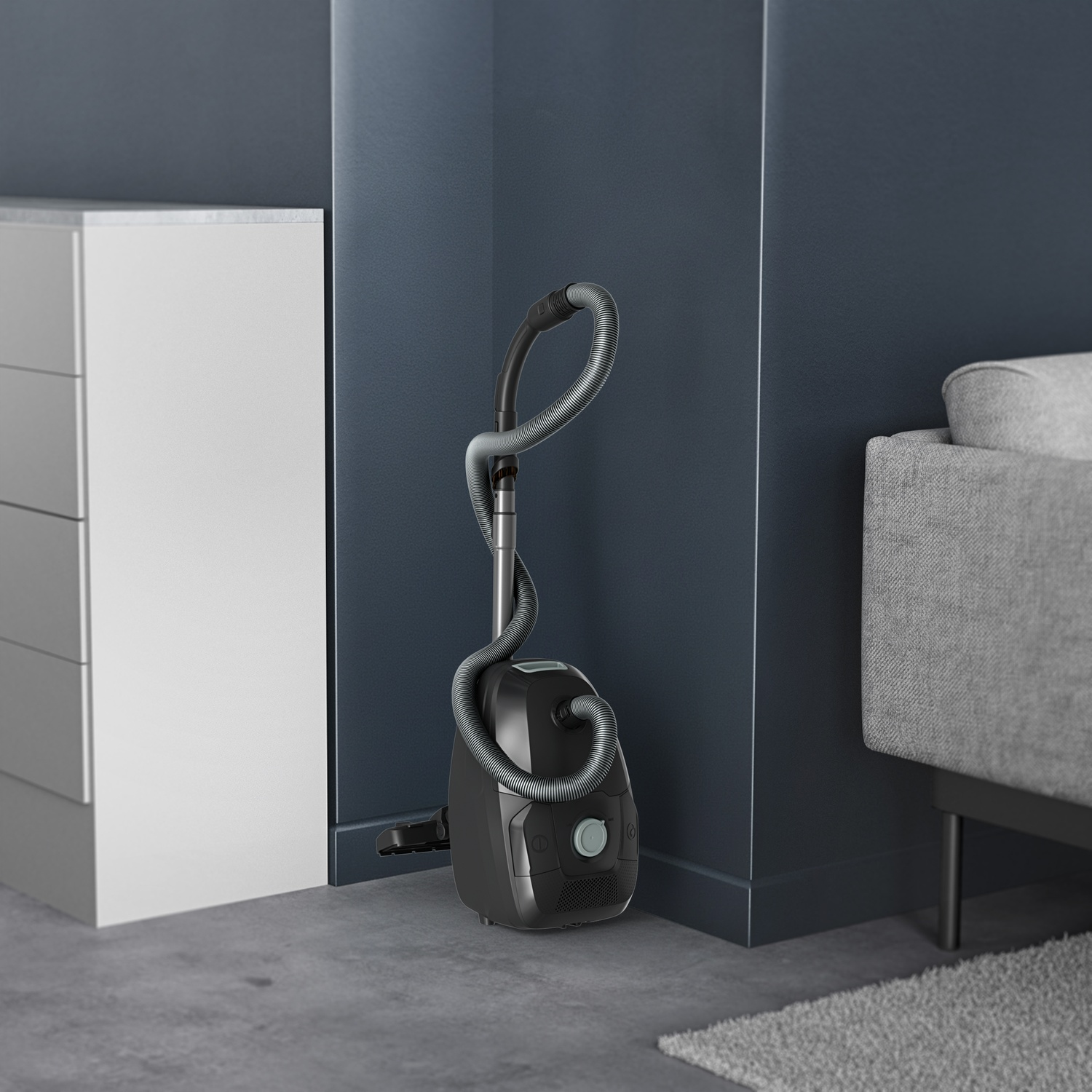 A gray vacuum cleaner stands on the floor in a corner between a white cabinet and a gray sofa.