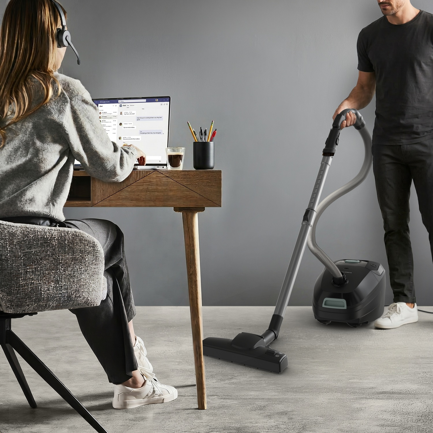 A woman works on a laptop at a desk while a man vacuums the floor with a black vacuum cleaner in the background.