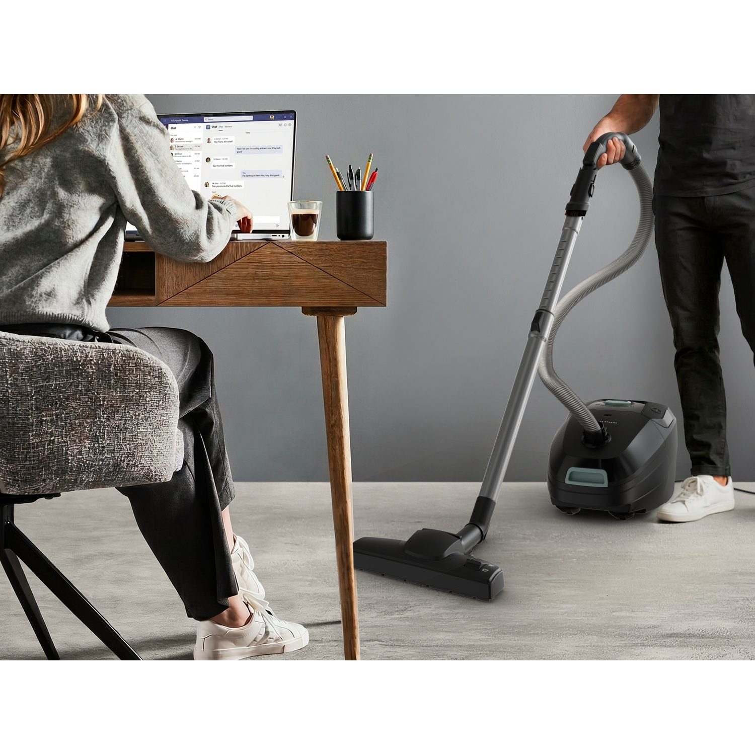 Woman sits at a desk working on a laptop, while a man in the background cleans the floor with a vacuum cleaner.