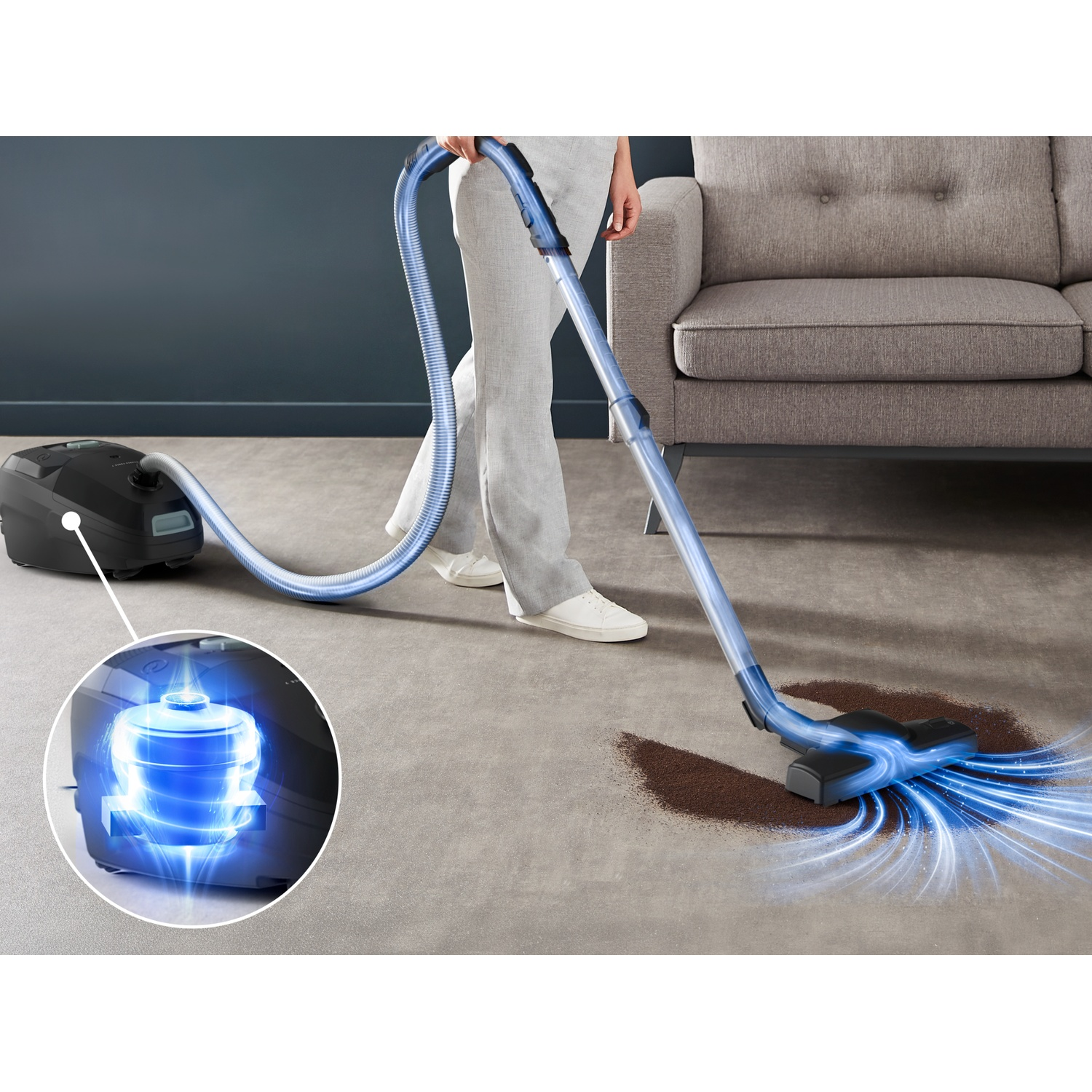 Person vacuuming brown dirt on carpet with a blue vacuum cleaner in front of a couch.
