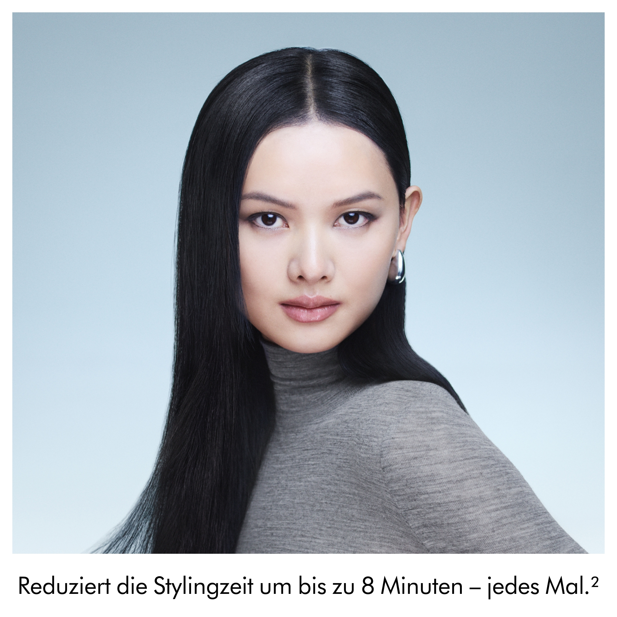 Eine Frau mit langem, glattem schwarzem Haar trägt einen grauen Rollkragenpullover vor hellblauem Hintergrund.