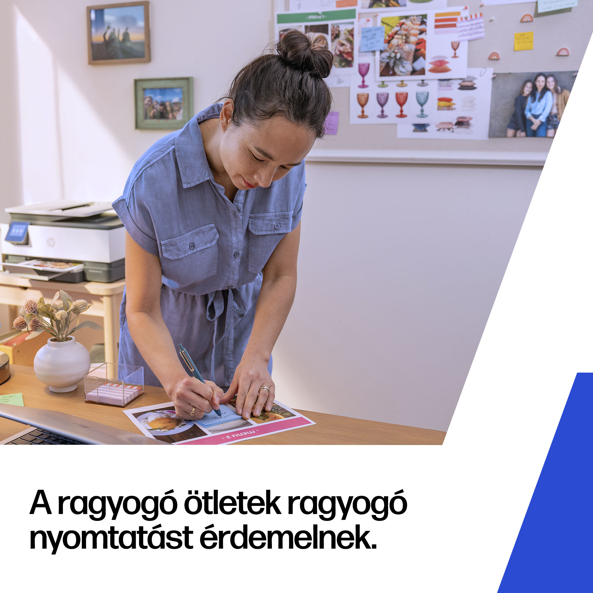 Egy kék inges nő papírra ír egy irodai asztalon, nyomtatóval és inspirációs táblával a háttérben.