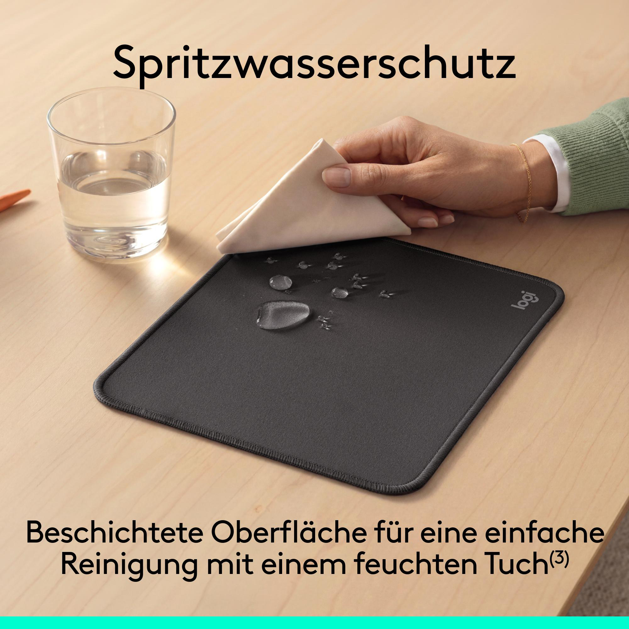 Eine Hand wischt Wassertropfen von einem schwarzen Mauspad neben einem Glas Wasser auf einem Holzschreibtisch.