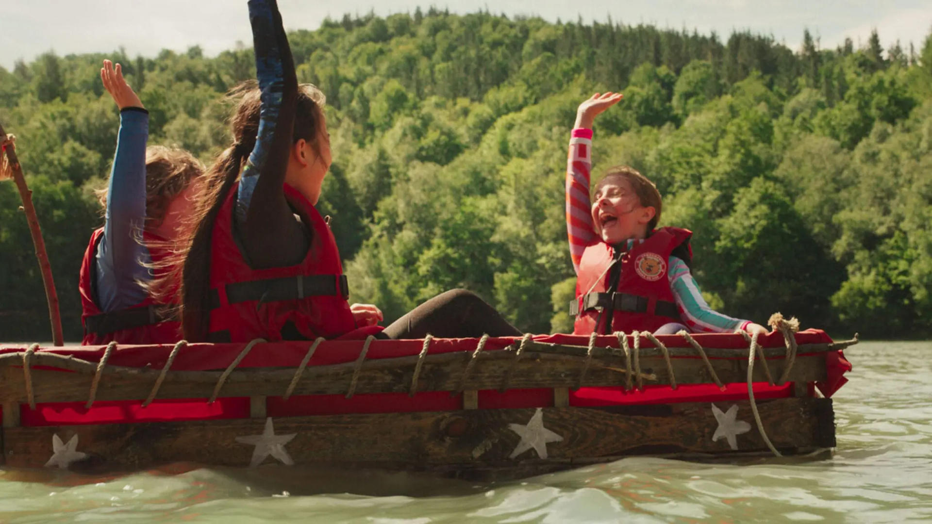 Drei Kinder in Rettungswesten winken fröhlich auf einem Holzfloß auf einem ruhigen See vor waldübersäten Hügeln.