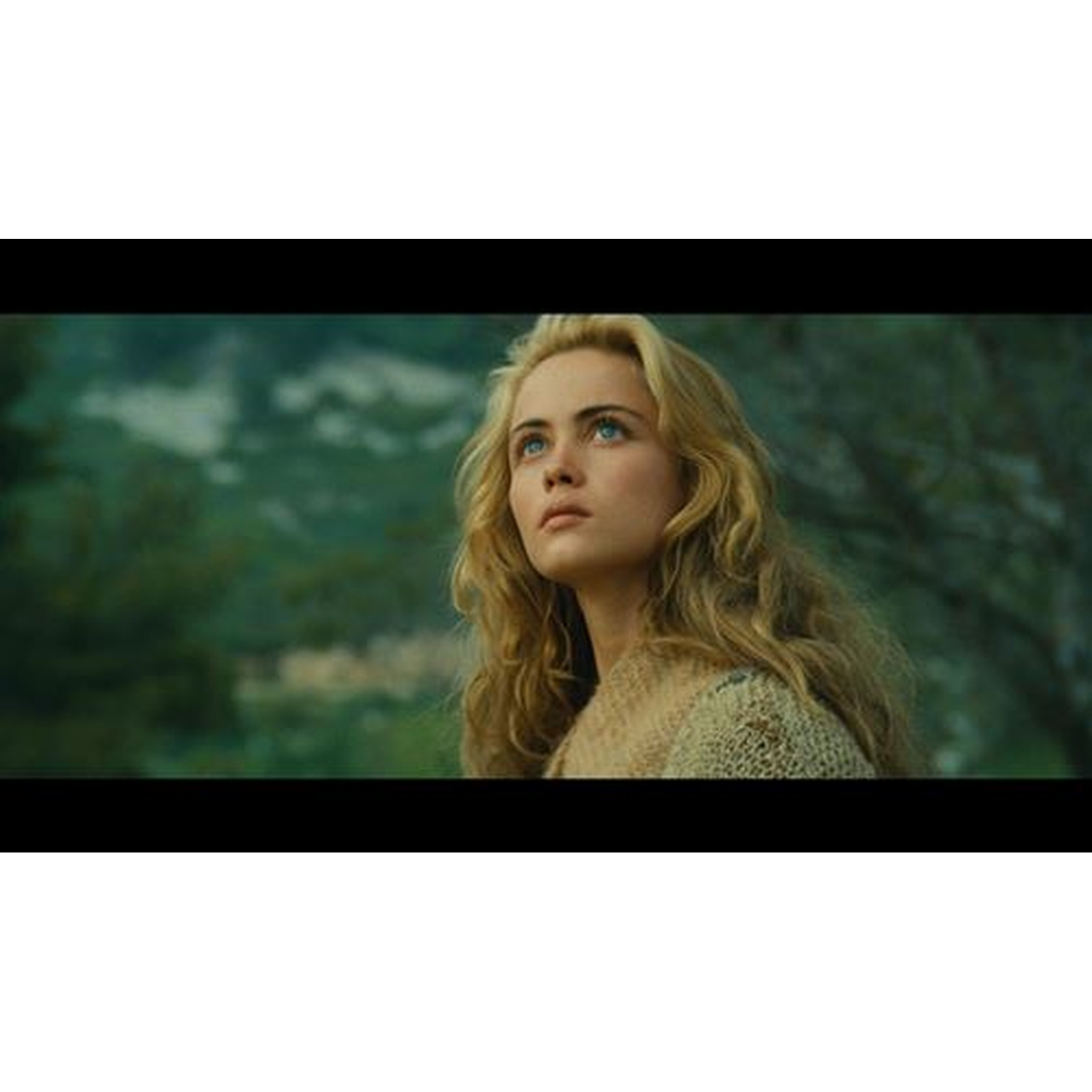 Une femme blonde aux yeux bleus regarde vers le haut devant un fond de forêt flou.