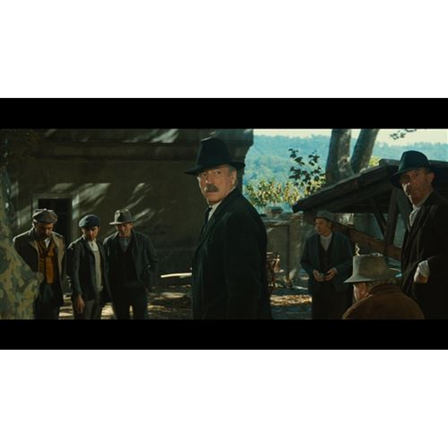 Groupe d'hommes en manteaux sombres et chapeaux debout à l'extérieur devant des bâtiments rustiques.