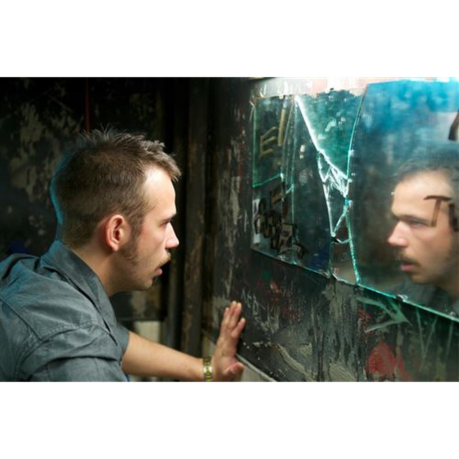 Een man met kort bruin haar kijkt in een gebroken, vuile spiegel in een donkere, met graffiti bedekte kamer.