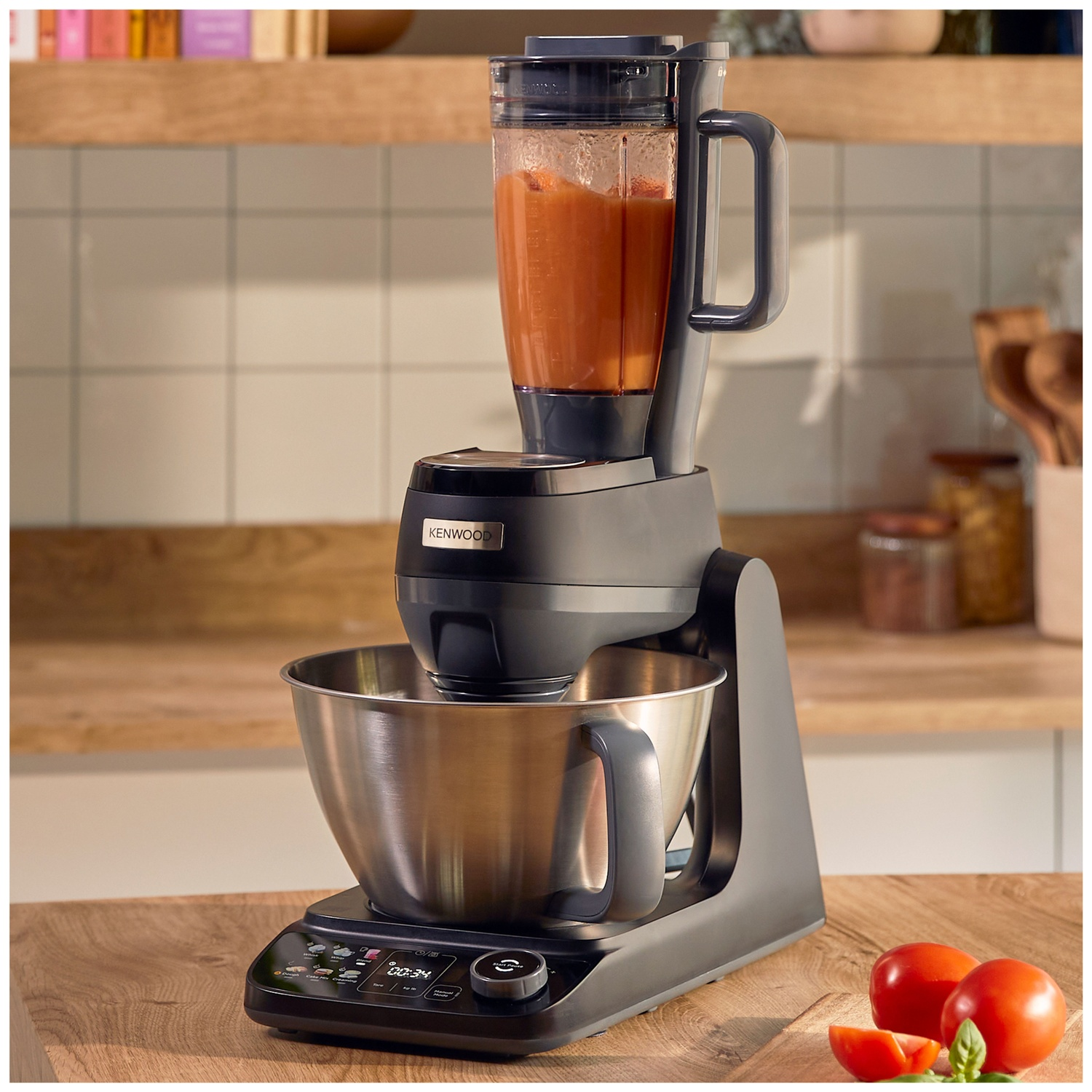 Een zwarte Kenwood keukenmachine met blender en oranje puree staat op een houten aanrecht met tomaten.