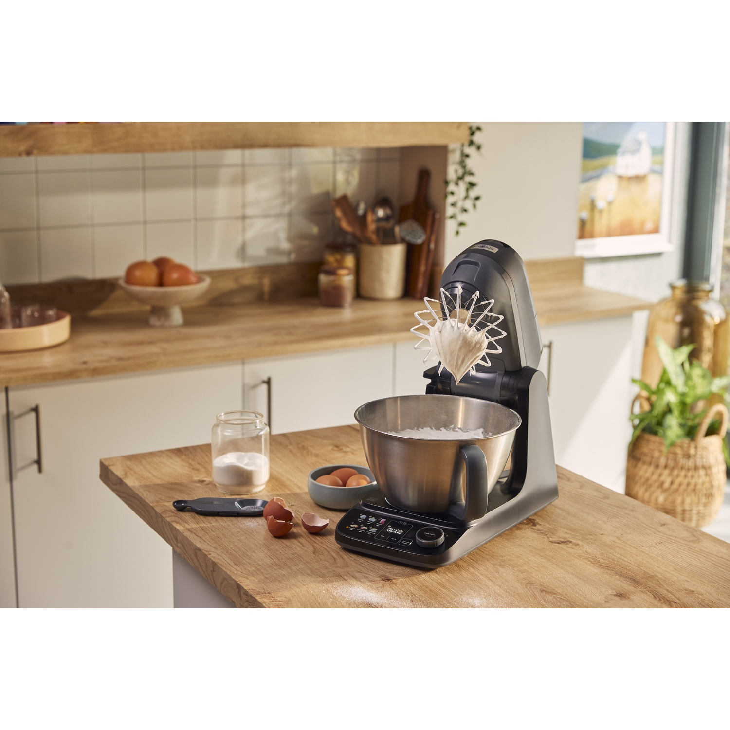 Een grijze keukenmachine op een houten aanrecht met kom, eieren en een pot suiker.