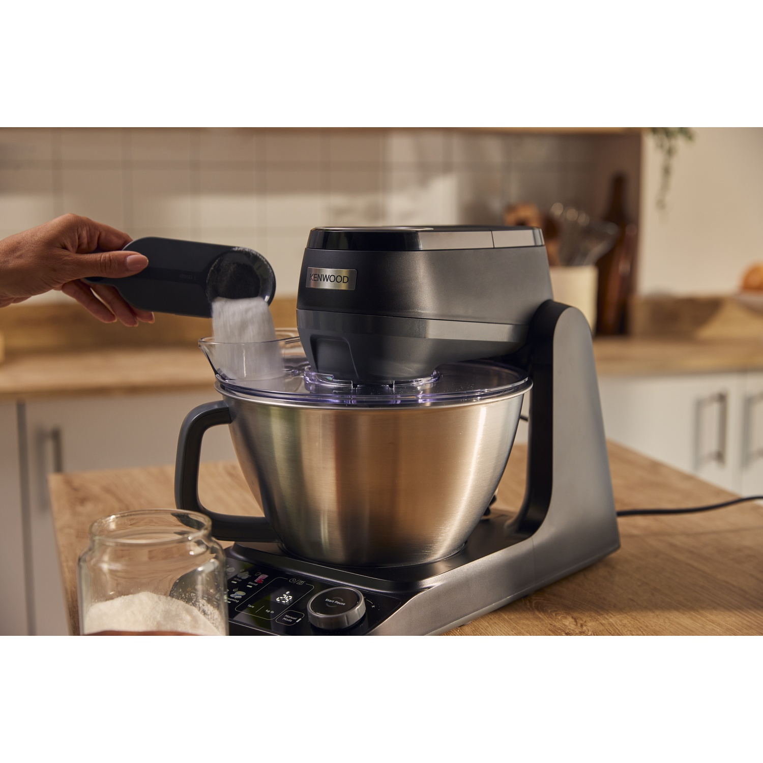 Een hand giet suiker uit een maatbeker in de roestvrijstalen kom van een zwarte Kenwood keukenmachine.