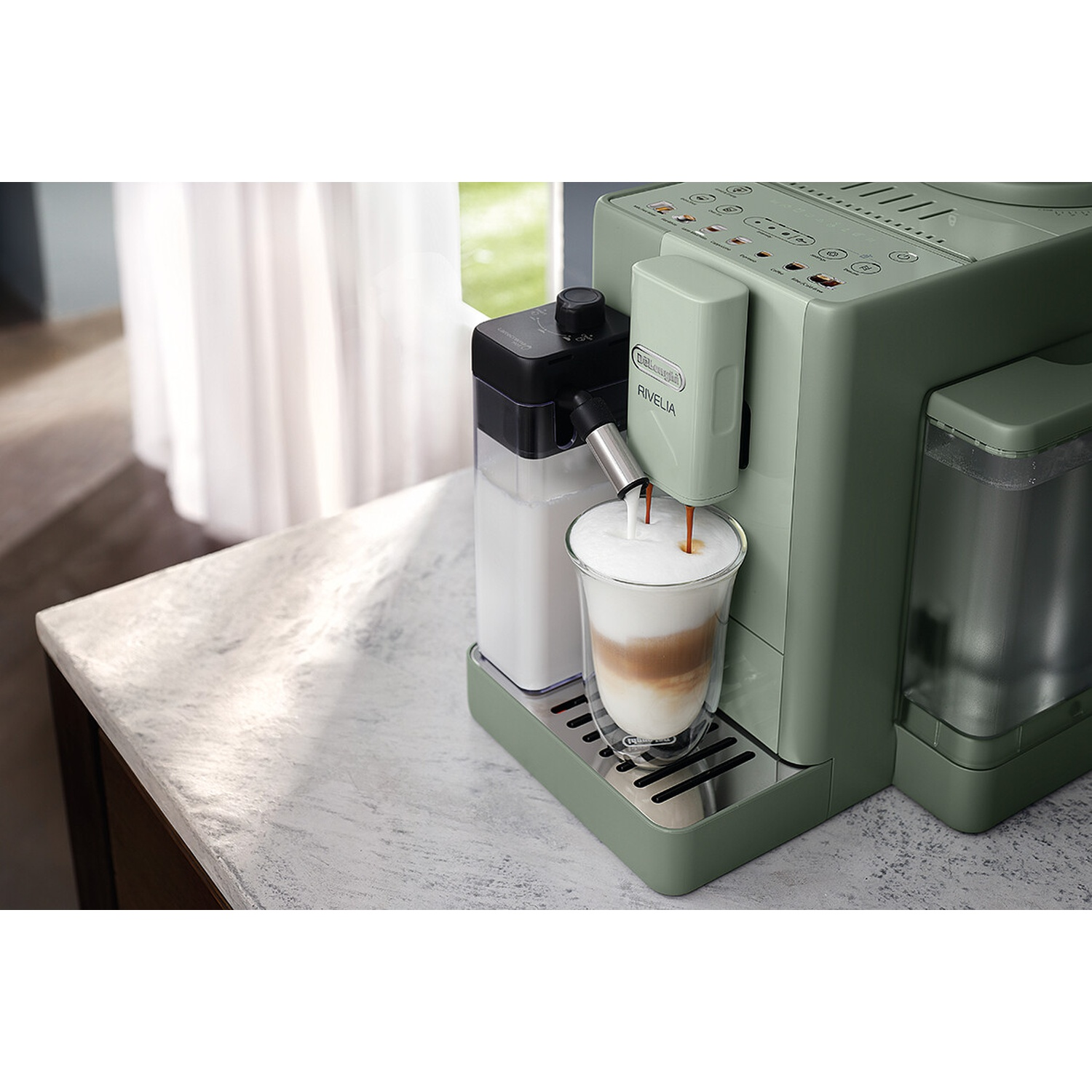 Een groene DeLonghi Rivelia koffiemachine schenkt melk en koffie in een glas op een lichtkleurig aanrecht.