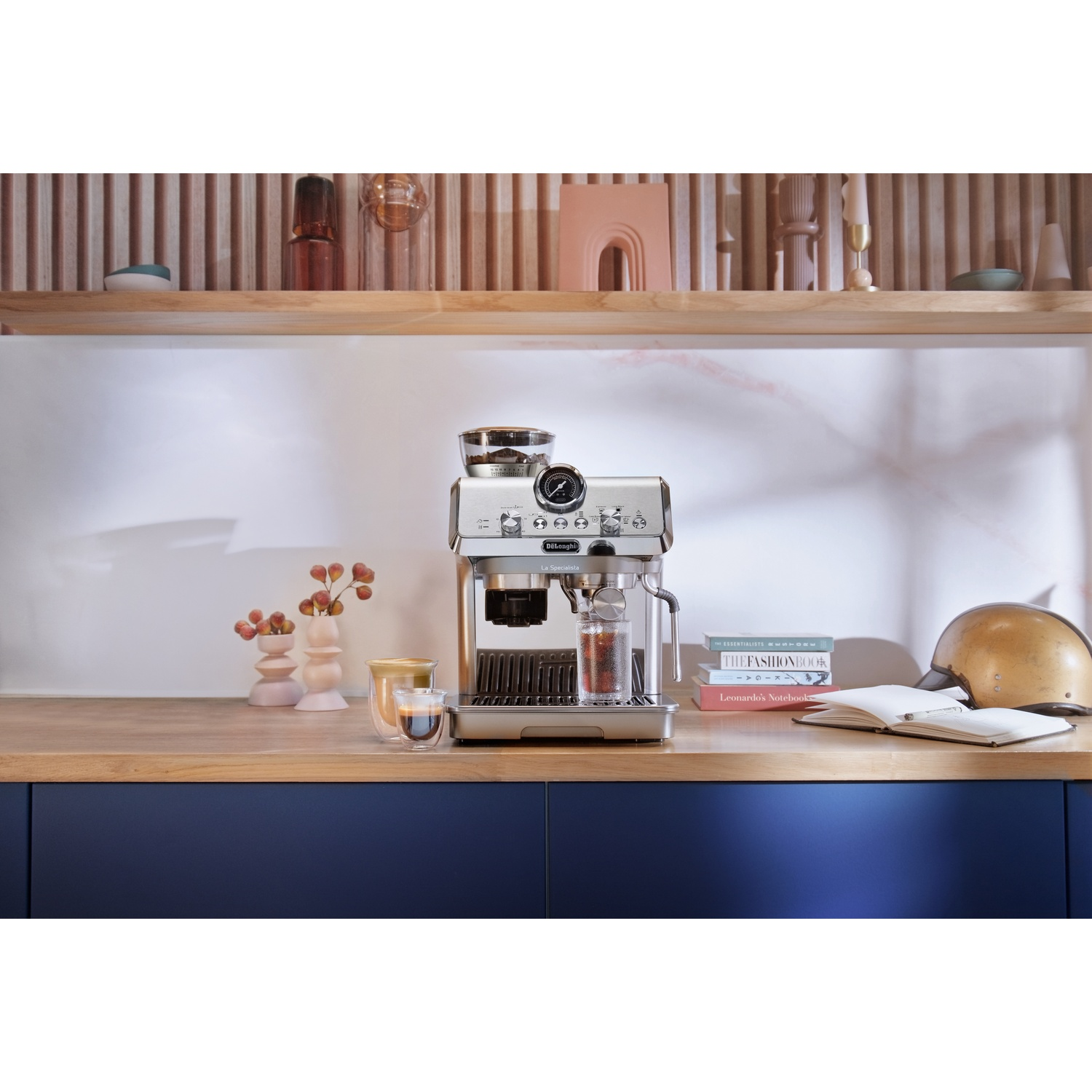 Een RVS koffiemachine op een aanrecht met boeken, een kopje koffie en een goudkleurige helm.