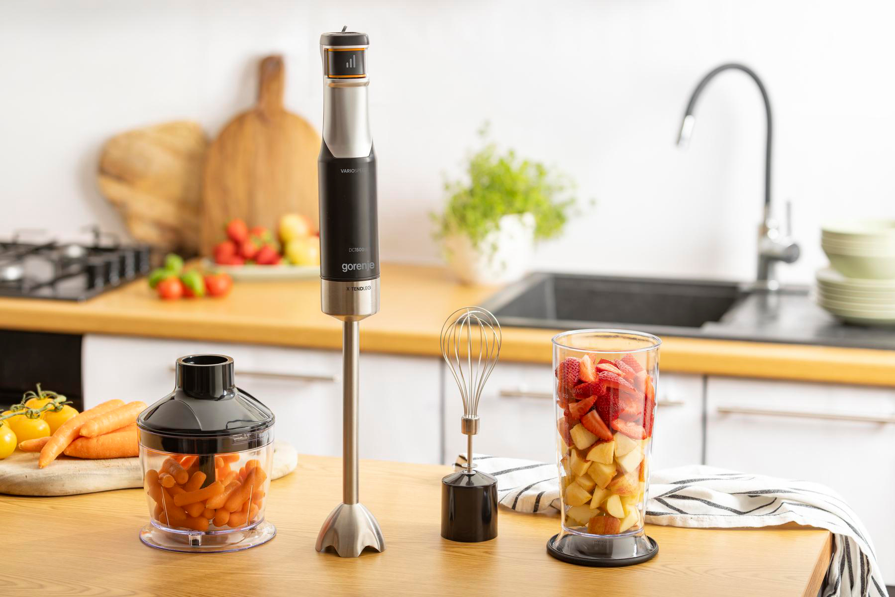 Edelstahl-Stabmixer mit Zubehör wie Zerkleinerer, Schneebesen und Mixbecher mit Erdbeeren auf einer Küchenarbeitsplatte.