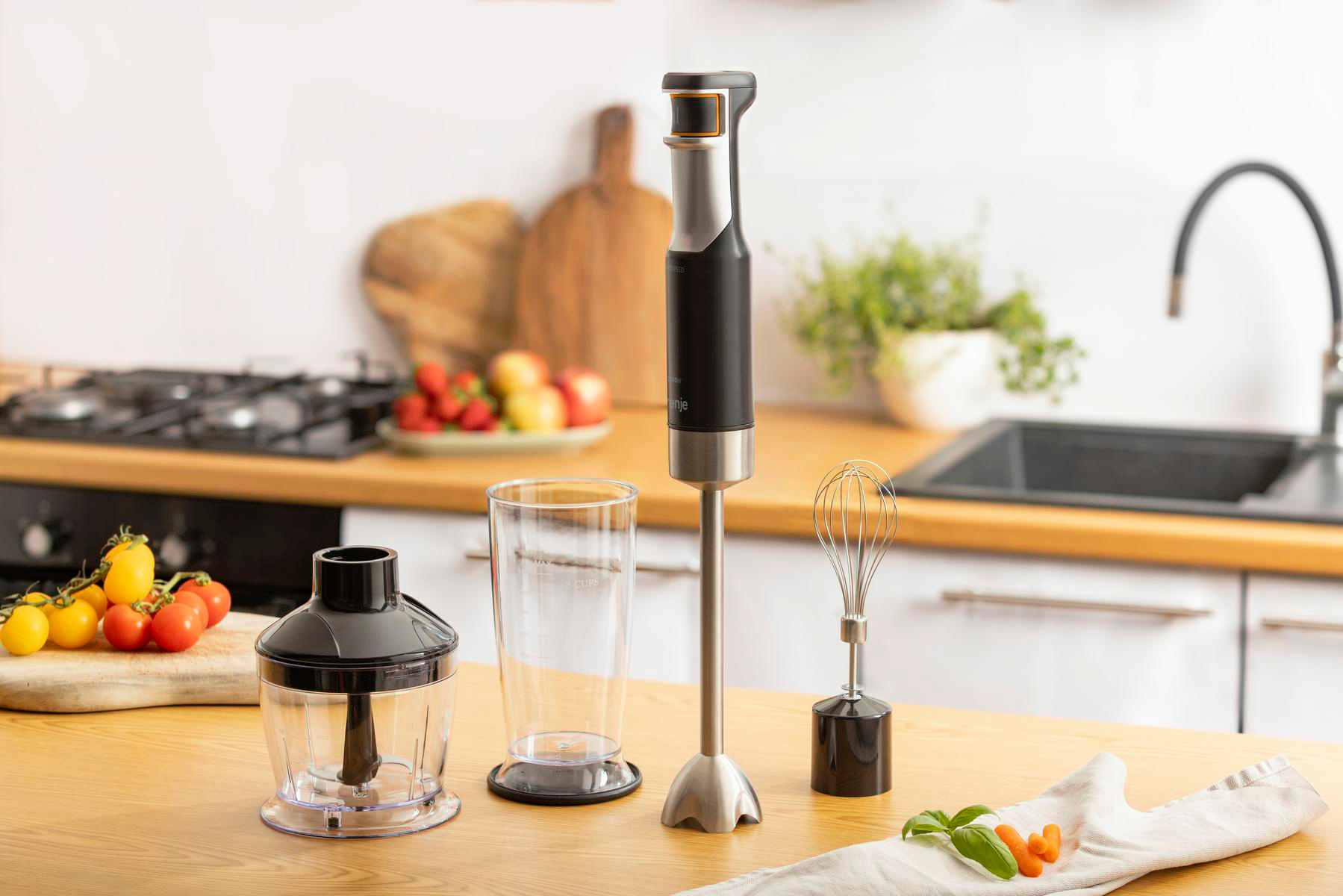 Ein schwarzer Stabmixer mit Zubehör und frischem Gemüse auf einer Holzarbeitsplatte in der Küche.