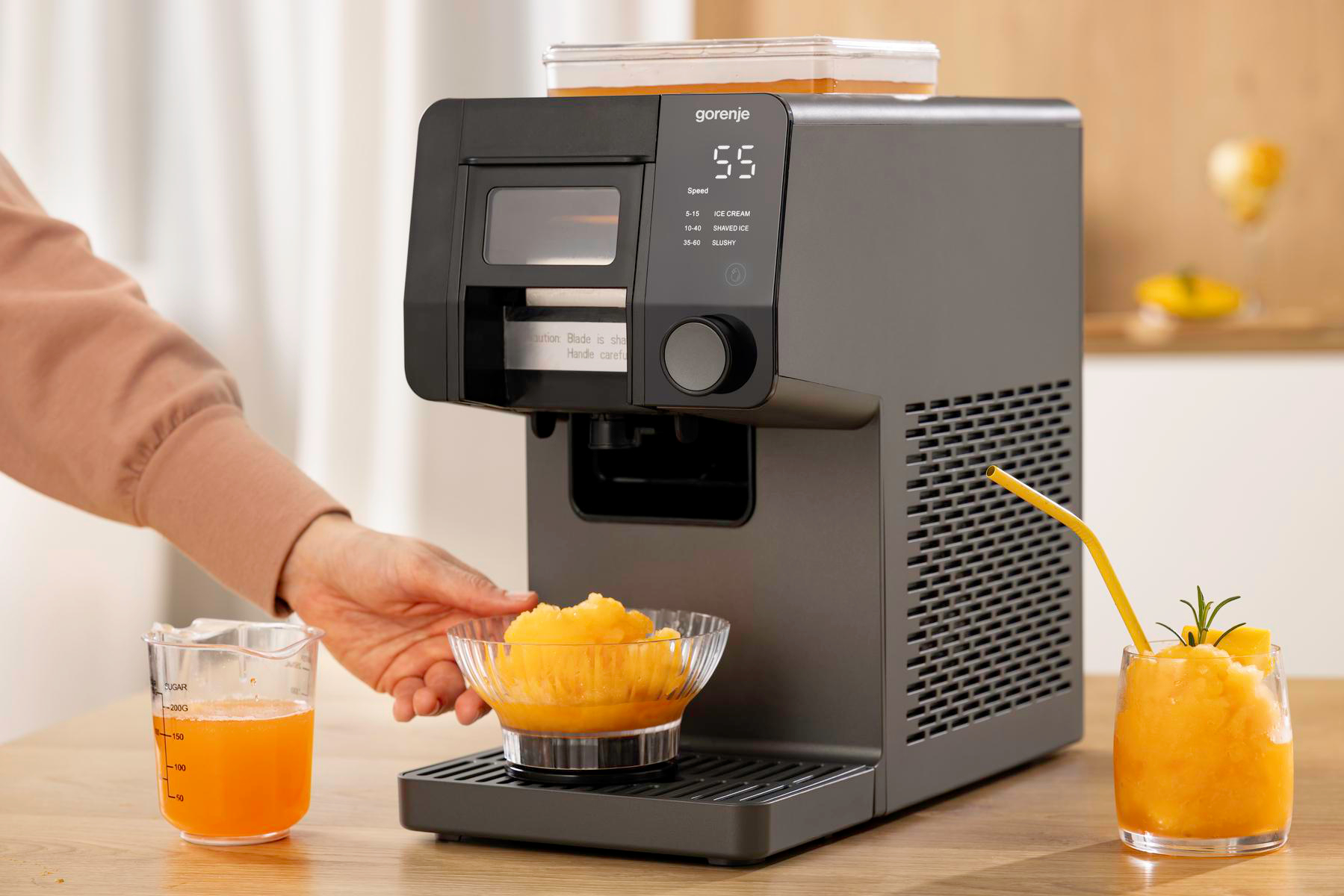 Eine schwarze Gorenje-Eishobelmaschine auf einer Theke, eine Hand hält eine Schüssel mit orangem Schabeeis.