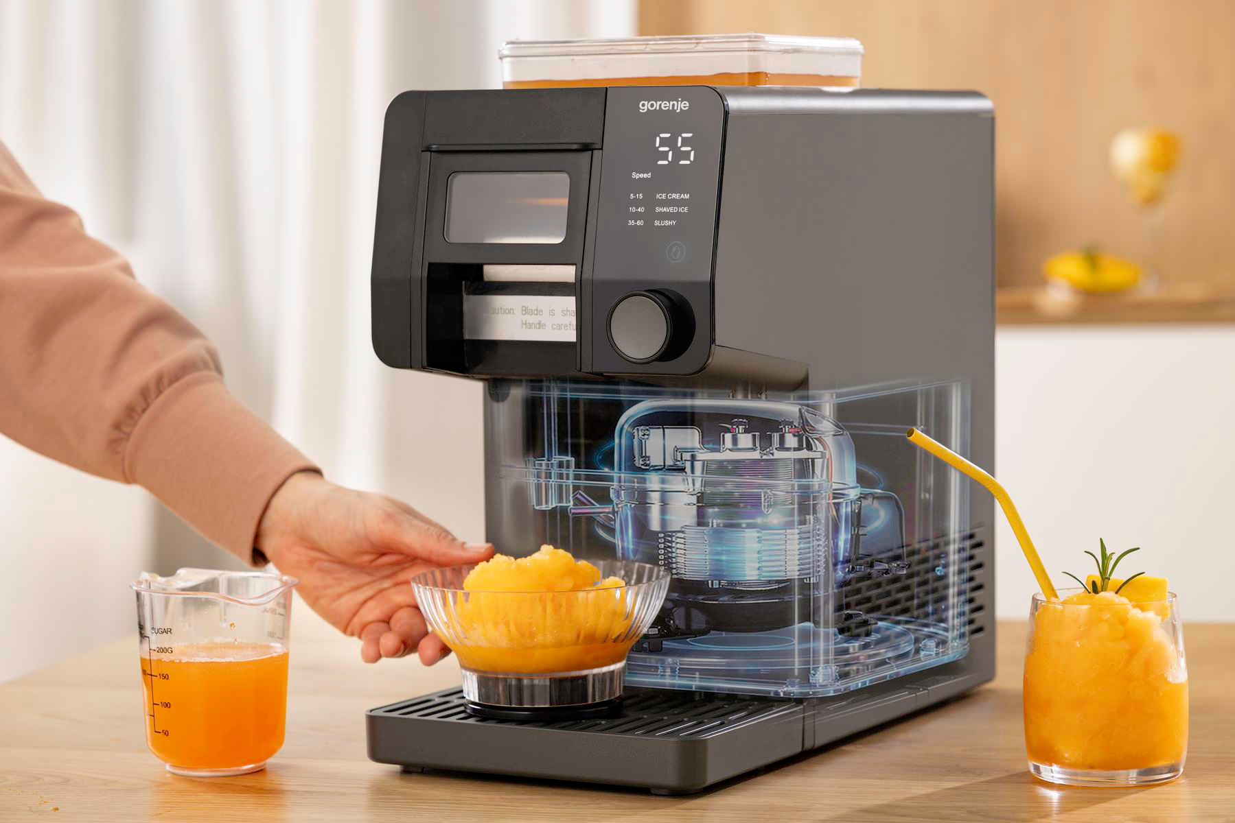 Schwarze Gorenje Eismaschine auf Holztisch, mit Glas Schabereis und Orangensaftbecher.