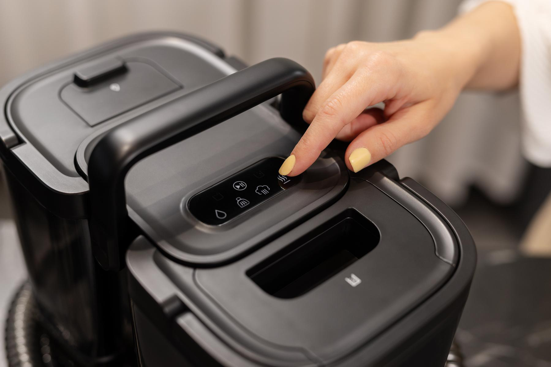 Eine Hand mit gelben Nägeln drückt einen Touch-Knopf auf einer modernen schwarzen Kaffeemaschine.