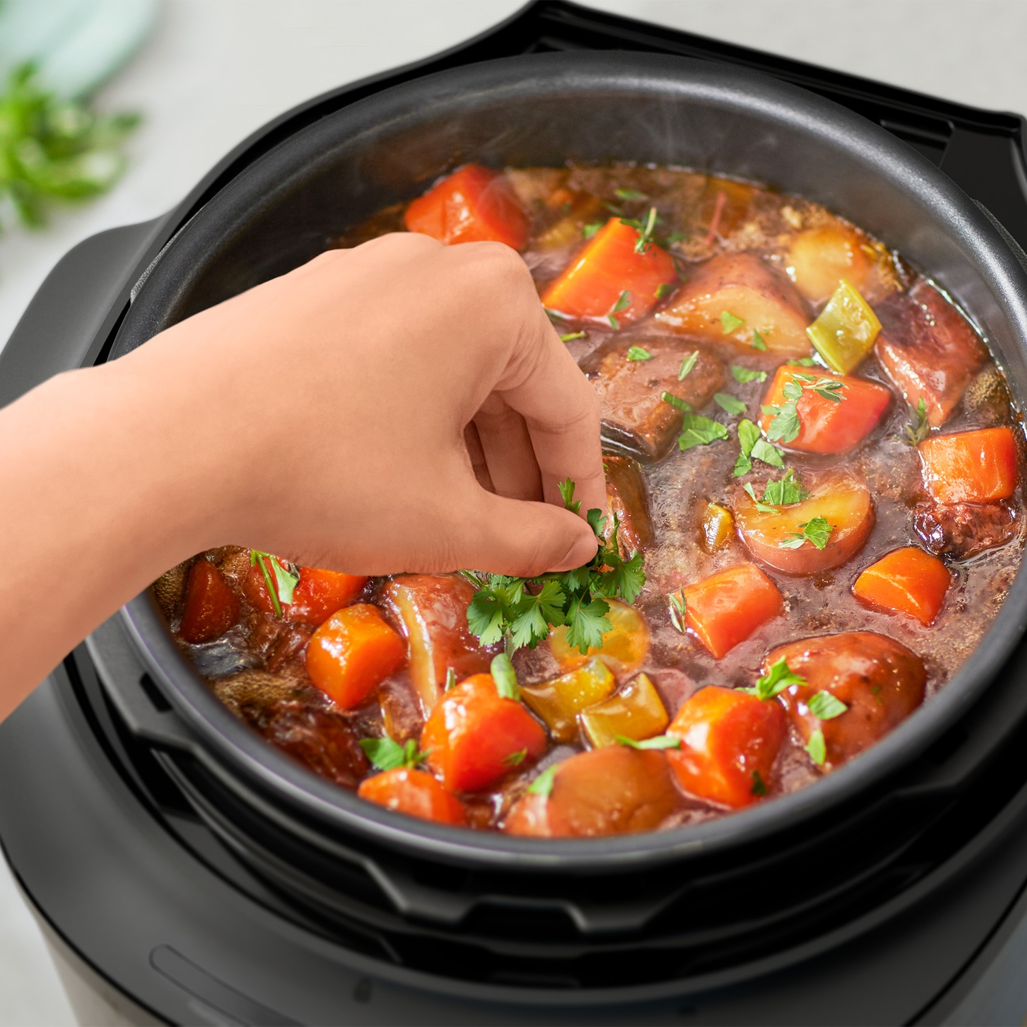 A hand adds fresh herbs to a steaming stew with potatoes and carrots inside a multicooker.