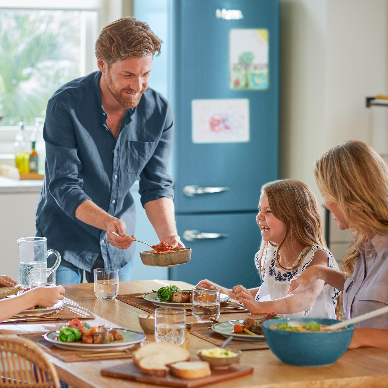 A father smiling while serving food to his family at a dining table in a kitchen.
