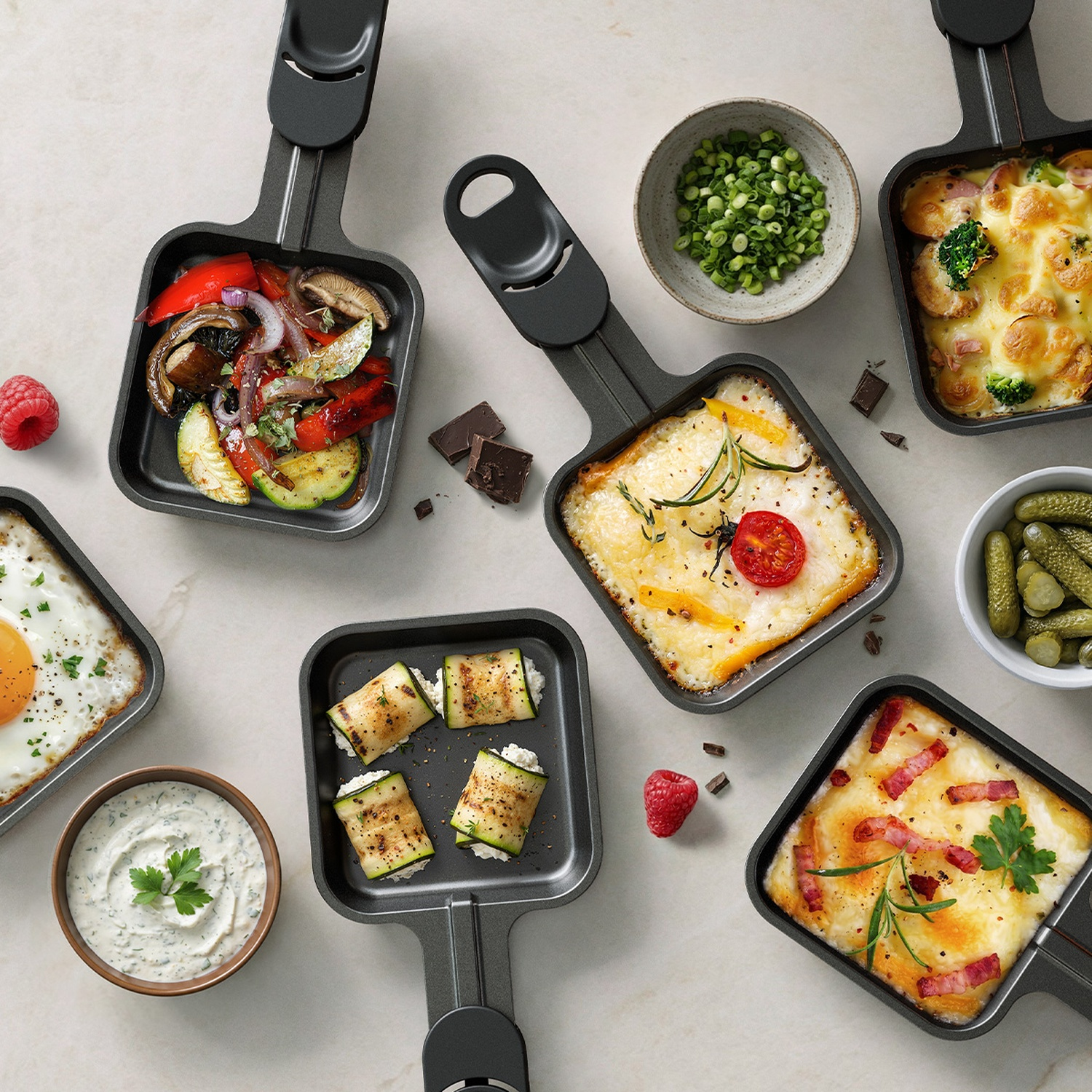 Raclette pans filled with vegetables, melted cheese, bacon, and dips on a light table.